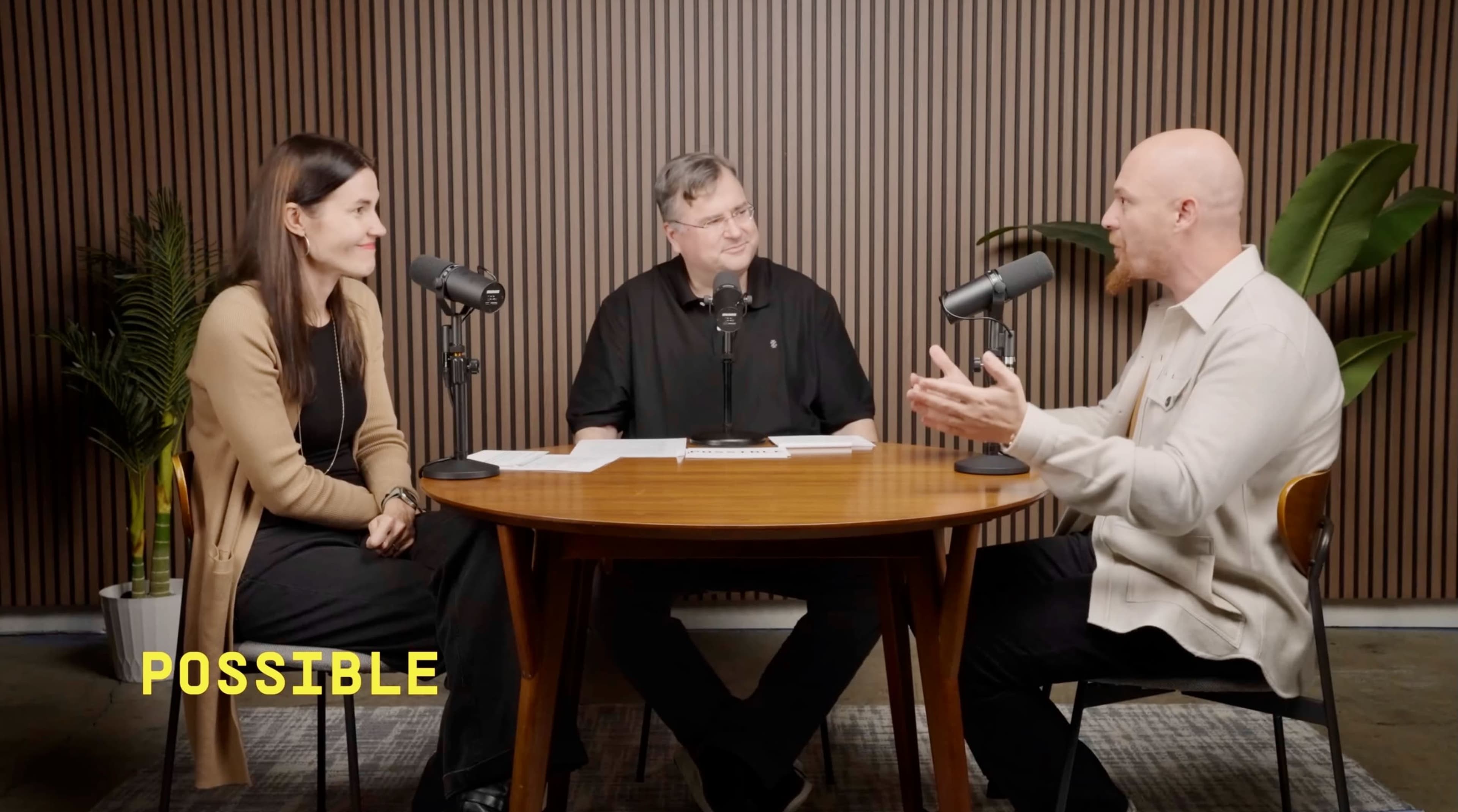 The image shows three people sitting around a wooden table with microphones, engaged in a discussion in a visually appealing interior.