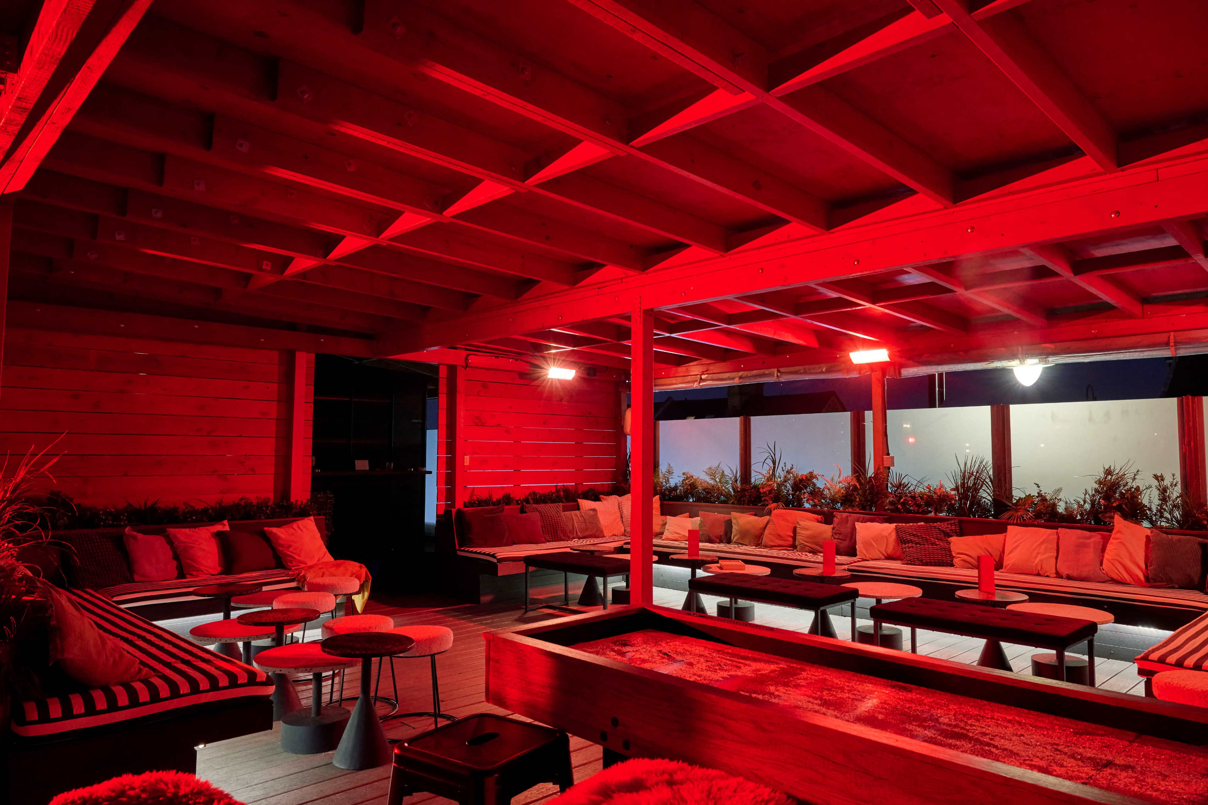The interior of a lounge is illuminated by red lighting, featuring wooden beams, comfortable seating, and small tables arranged throughout the space.