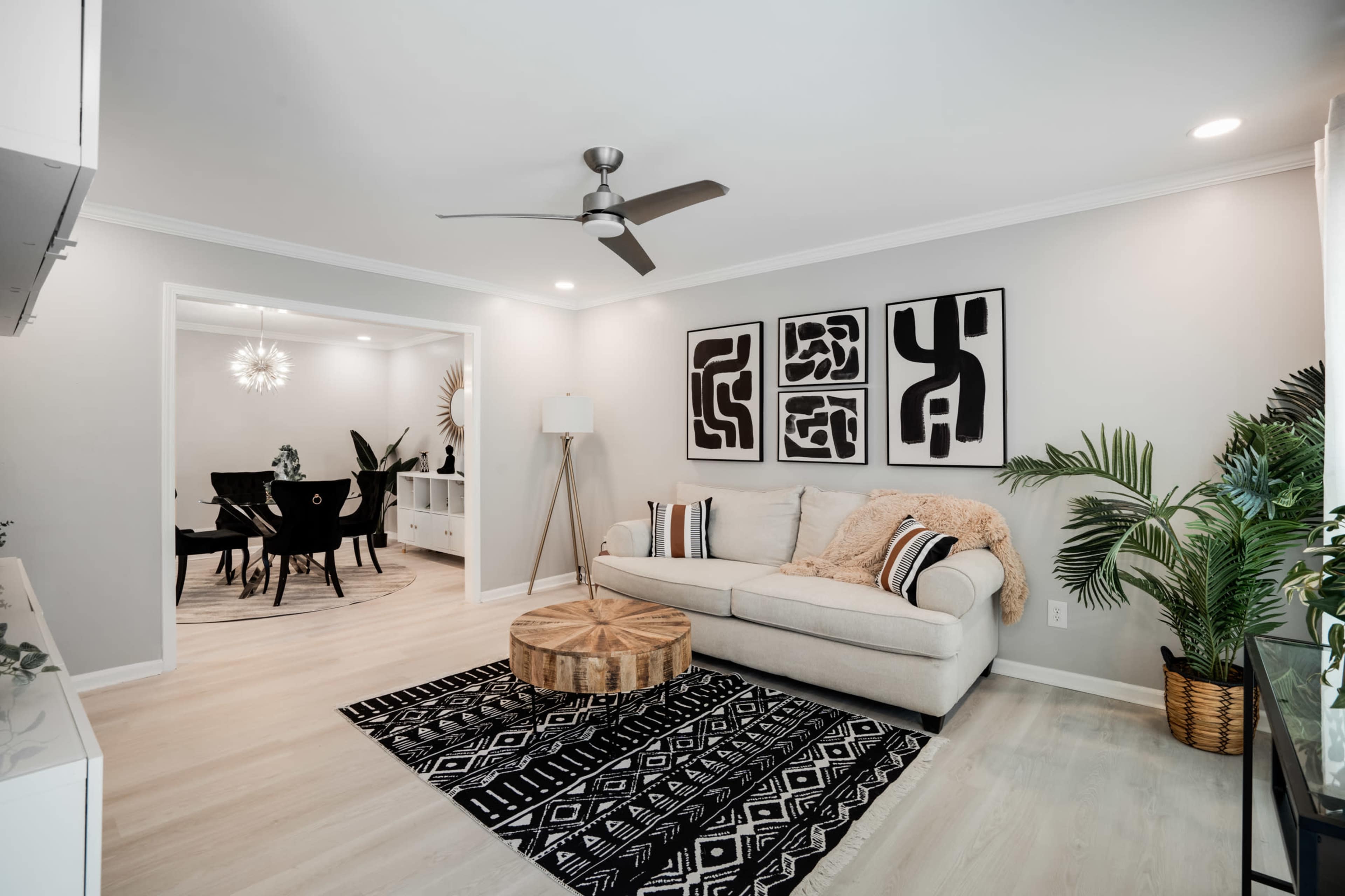 A modern living room features a light-colored sofa, a patterned rug, and framed artwork on the walls, with a view of a dining area in the background.