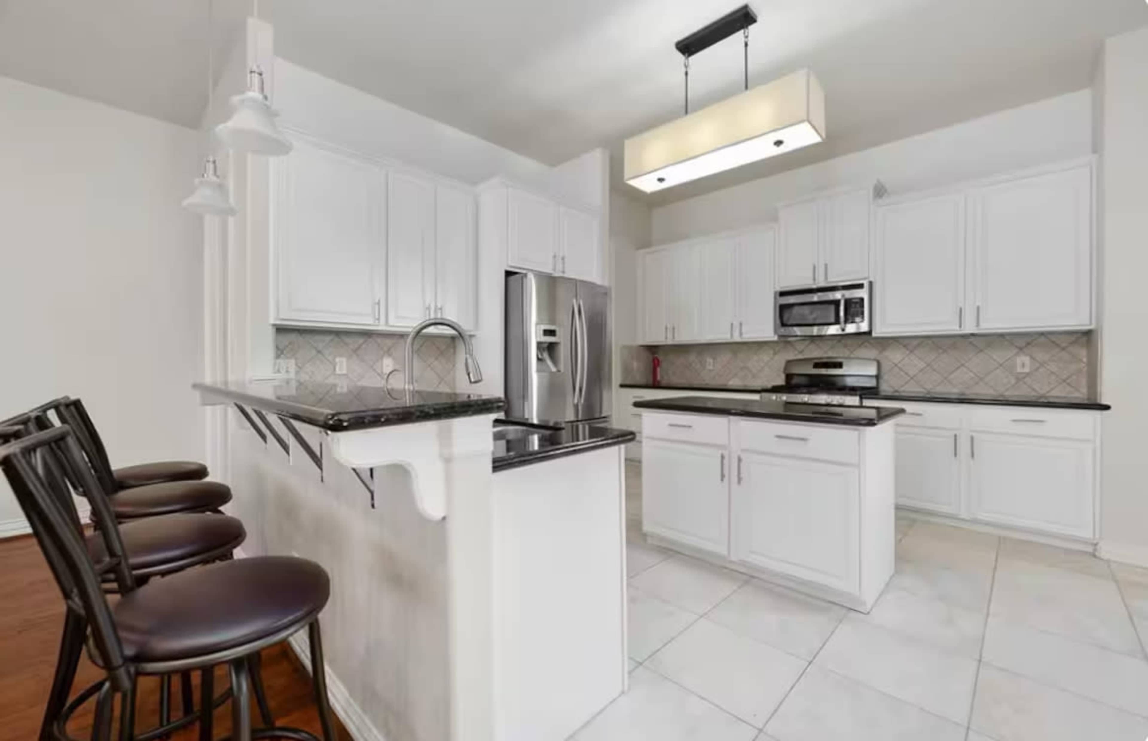 The image shows a modern kitchen with white cabinetry, stainless steel appliances, and a countertop that extends into a bar with seating.