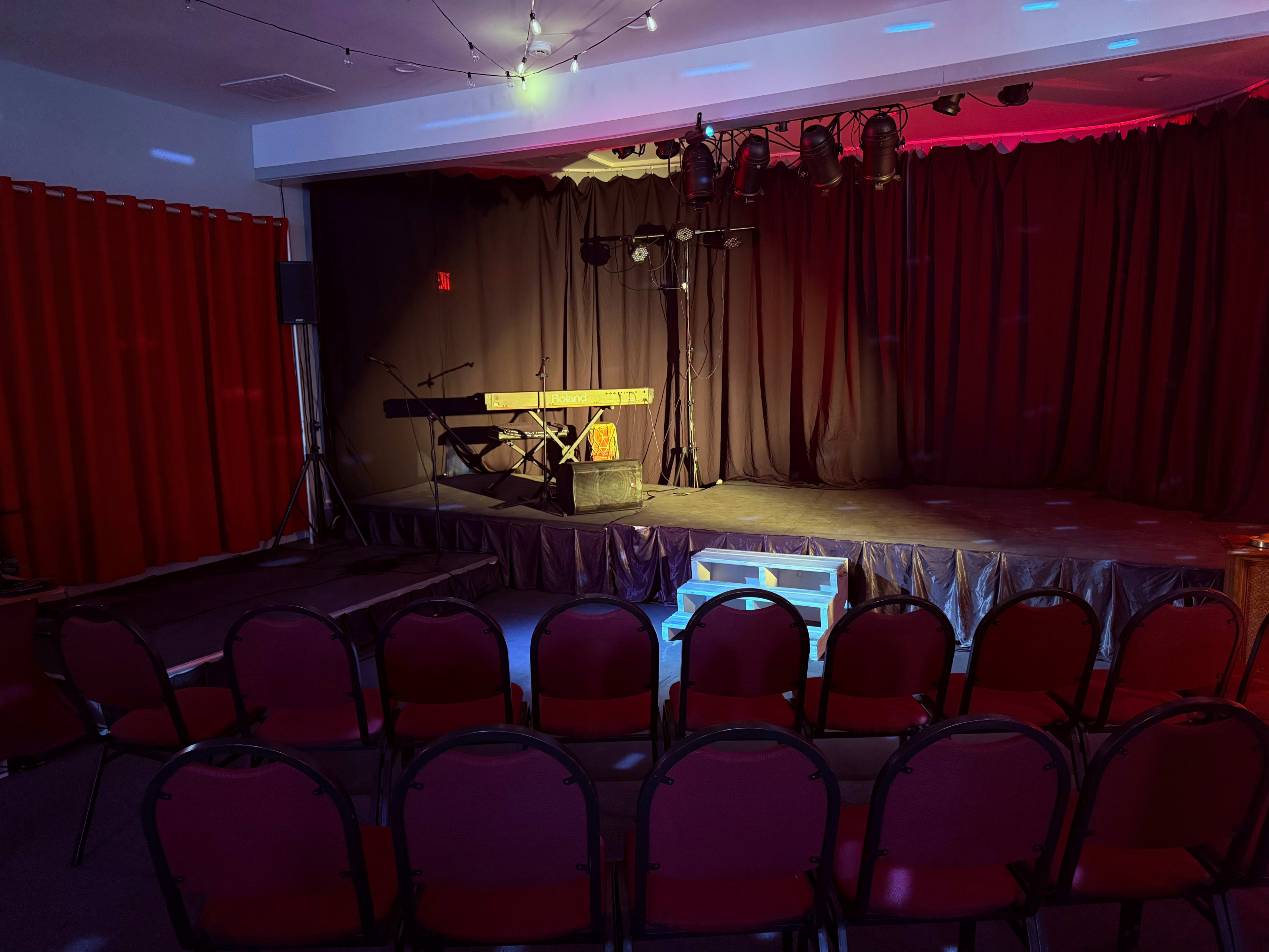 The image shows a small stage with a keyboard and microphone, surrounded by red curtains and empty red chairs arranged in front.