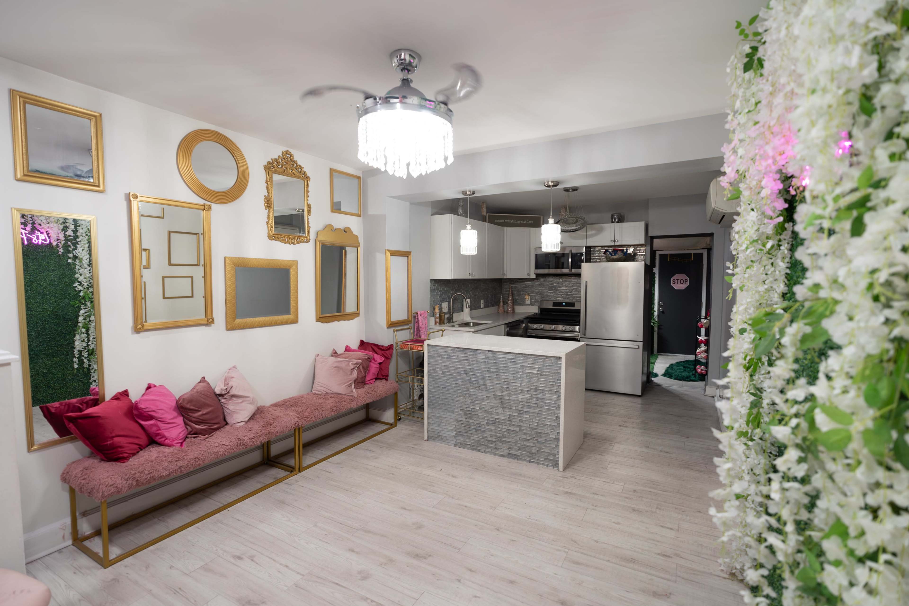 The image depicts a modern, stylish interior featuring a kitchen with stainless steel appliances, a seating area with decorative pillows, and a wall adorned with greenery and flowers.