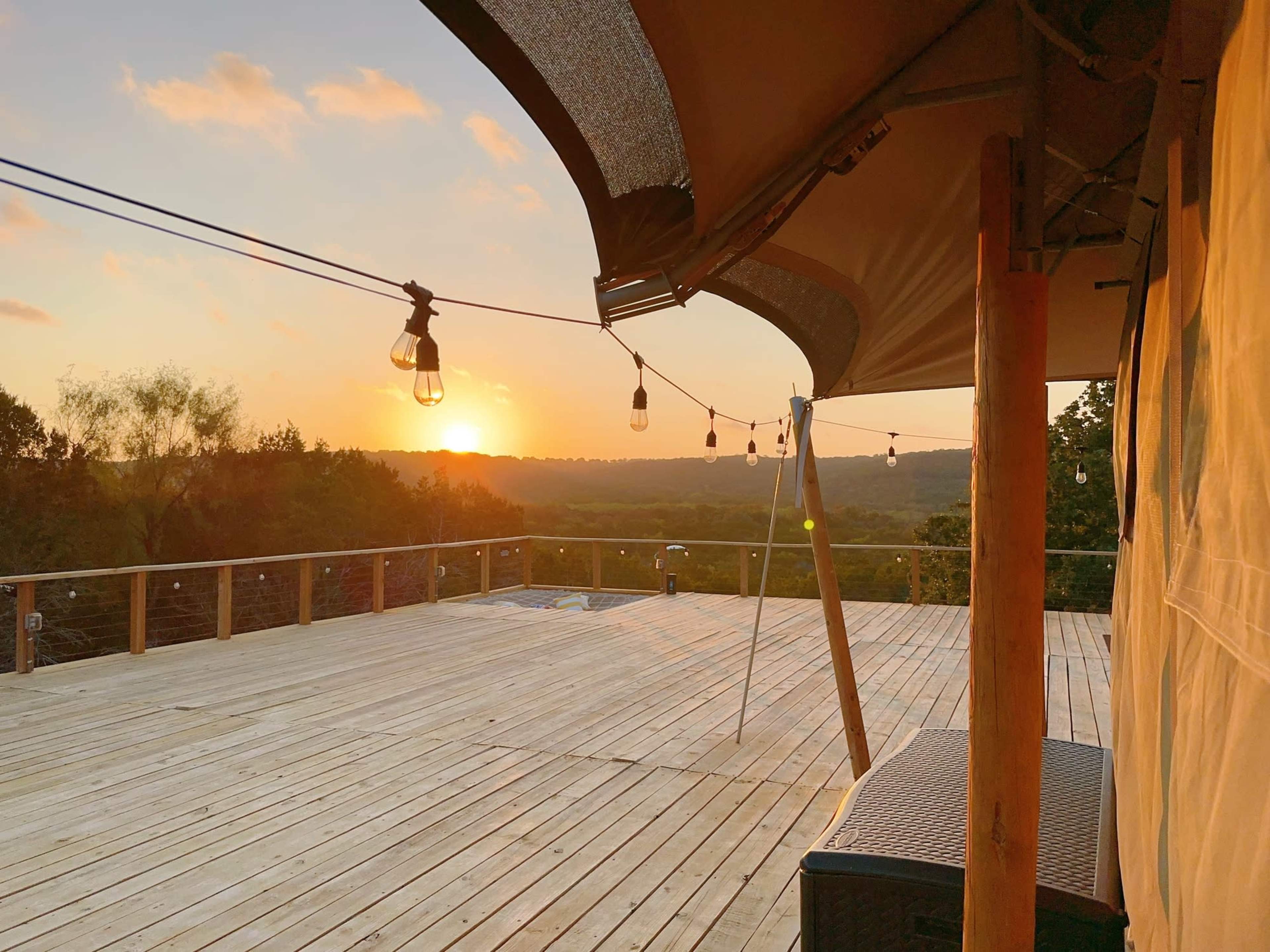 A wooden deck with string lights overlooks a sunset and rolling hills in the distance.