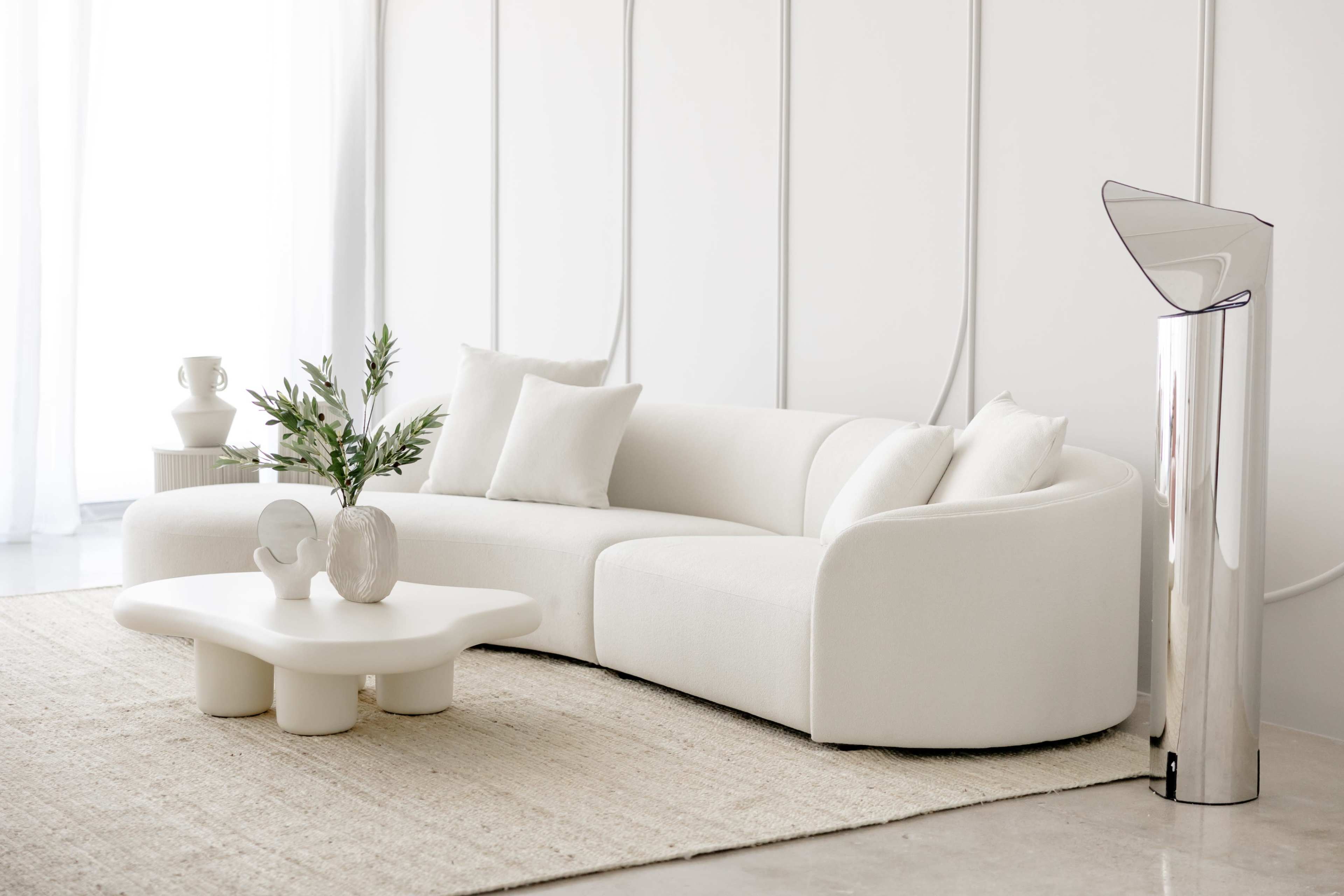 The image features a modern, white sectional sofa with decorative pillows, arranged next to a round coffee table on a textured rug, with a vase and a decorative object displayed on the table.