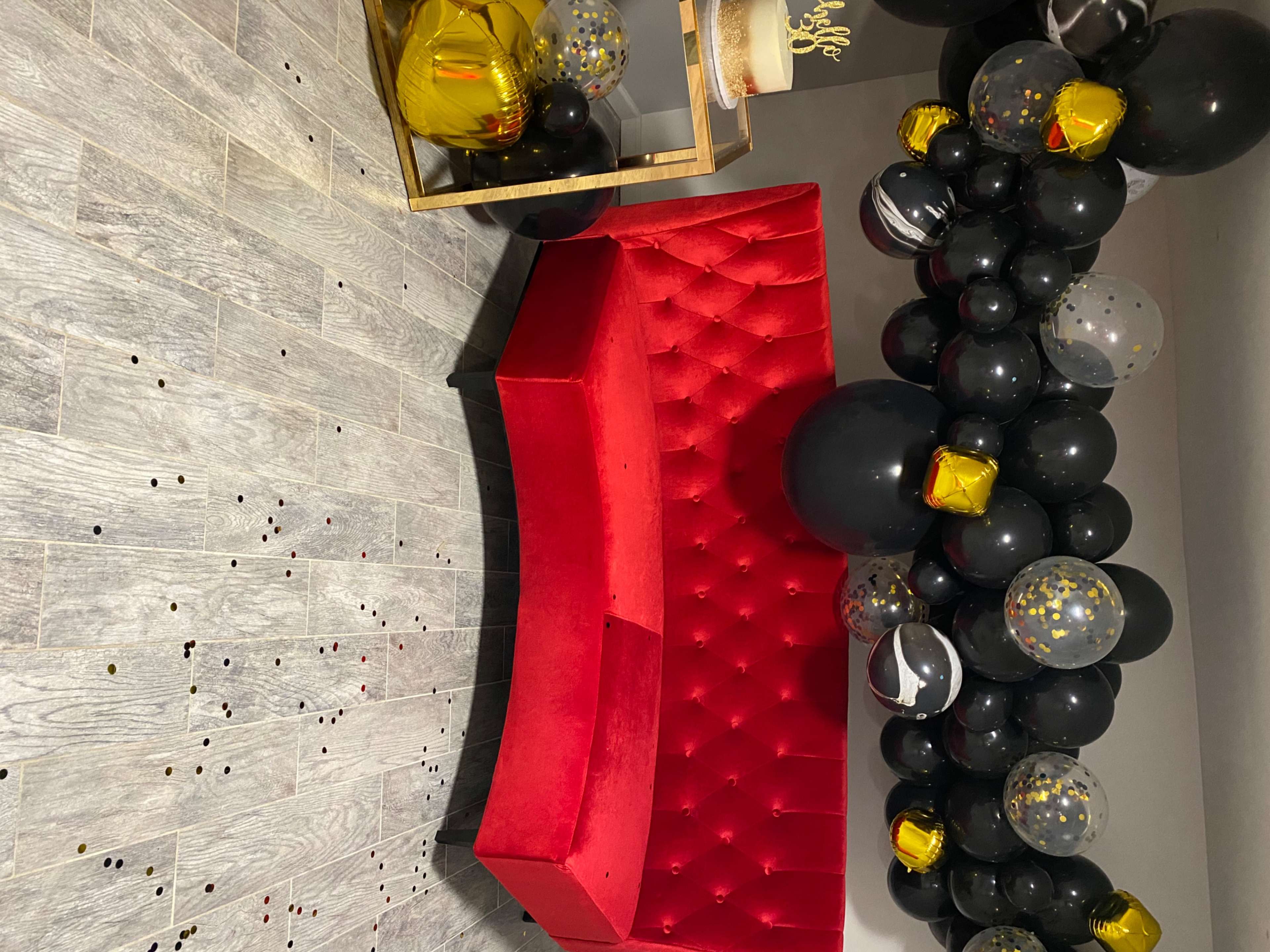 A red tufted sofa positioned near a decorative table adorned with black and gold balloons, set against a gray tiled floor sprinkled with confetti.
