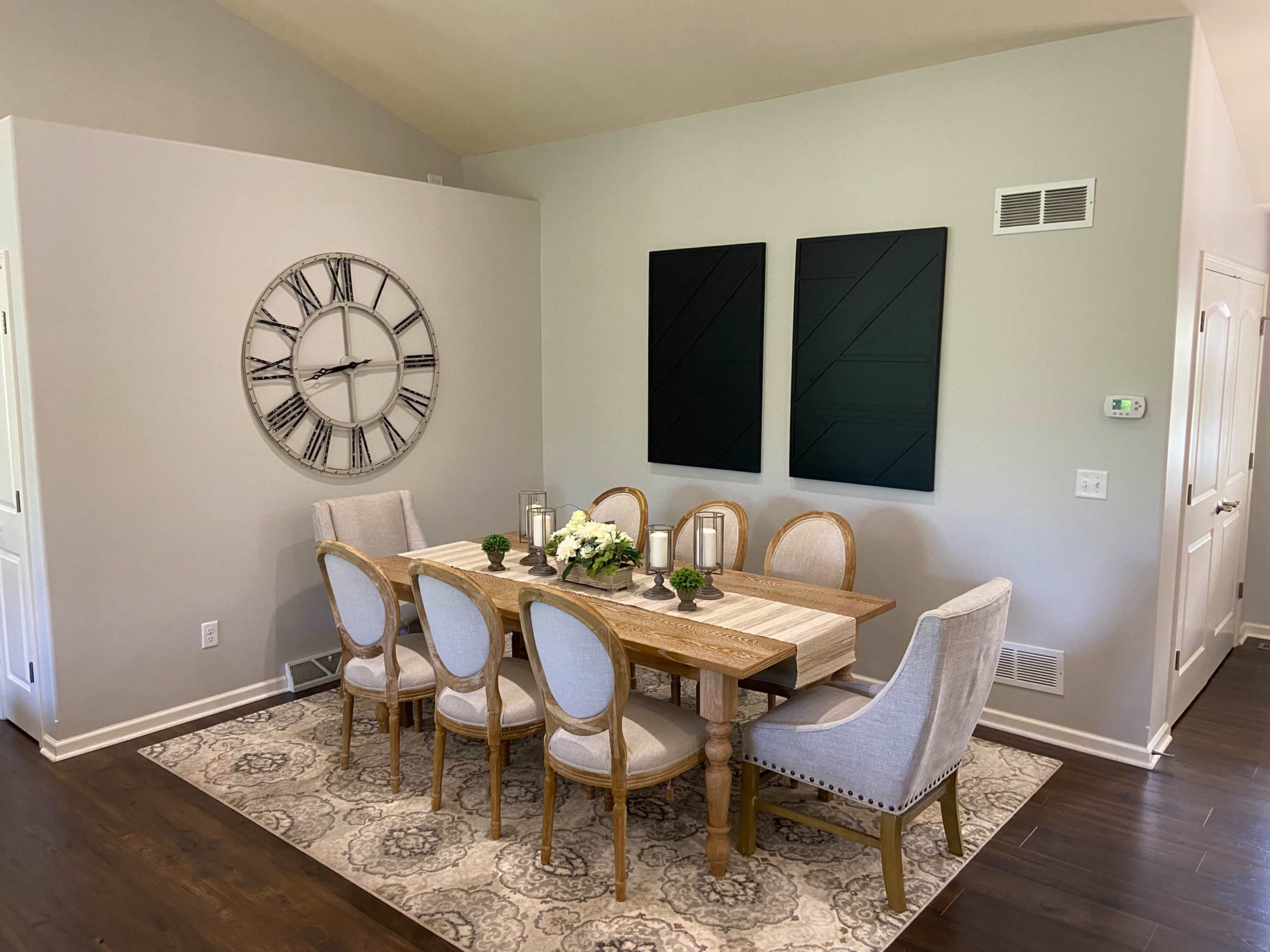 A dining room features a wooden table surrounded by eight chairs, with a large wall clock and two black wall art pieces.