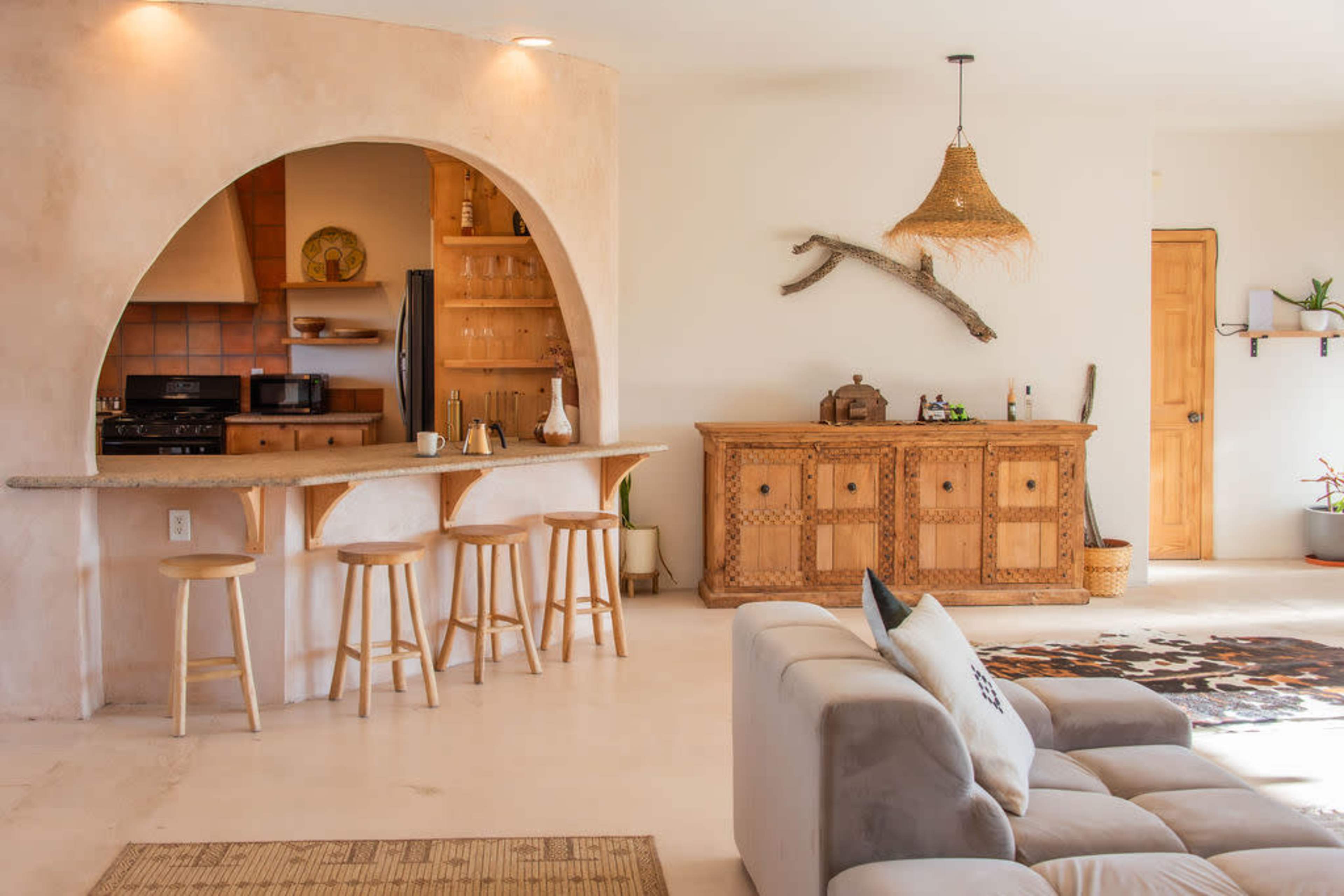 The image depicts a modern, open-concept living space featuring a kitchen bar with three stools, a wooden sideboard, and a cozy sofa, all complemented by natural light and minimalist decor.