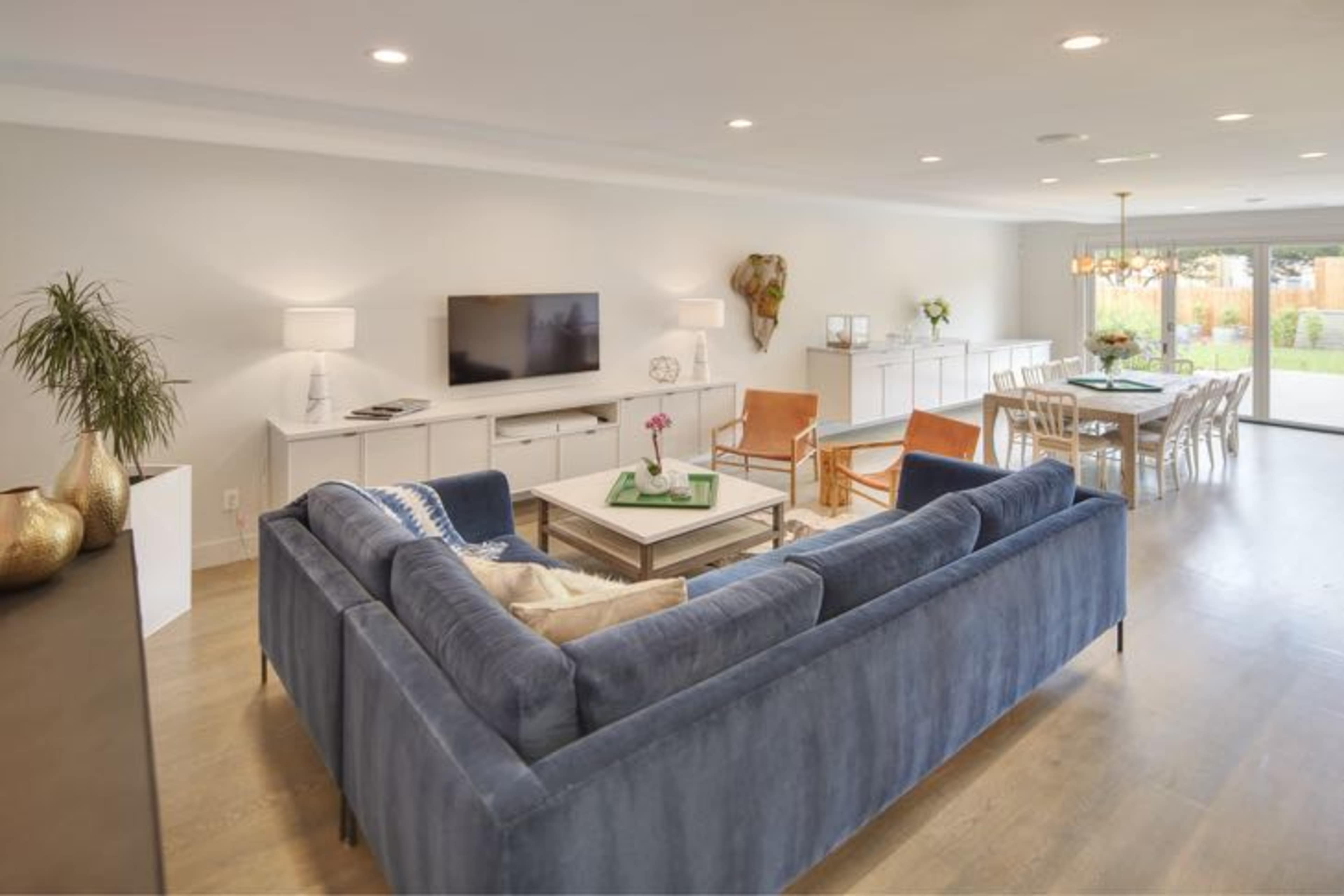 A modern living room with a blue sectional sofa, a coffee table, a television on a white media console, and a dining area with wooden chairs in the background.