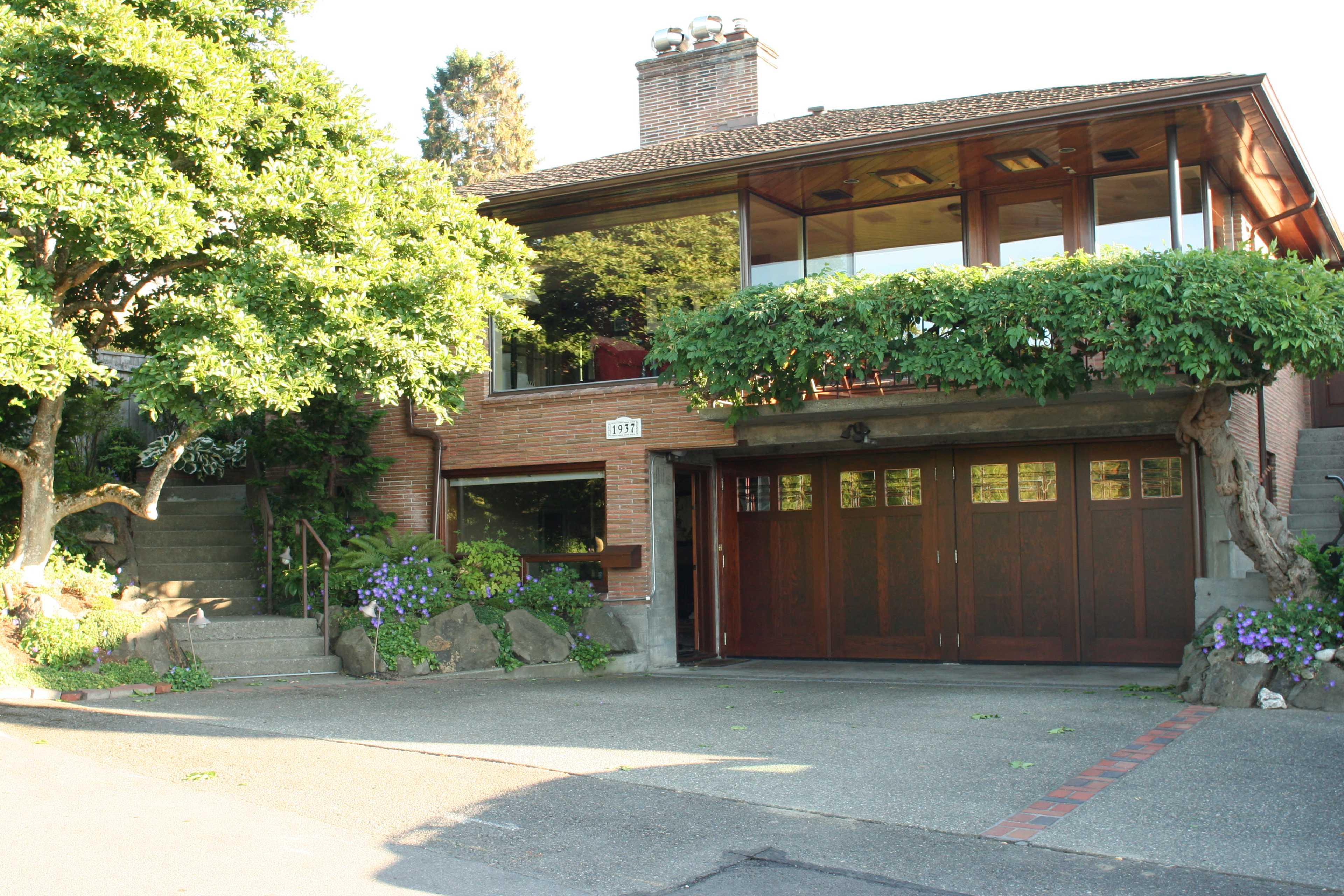 A modern two-story house with large windows and a wooden garage door, surrounded by greenery and steps leading to the entrance.