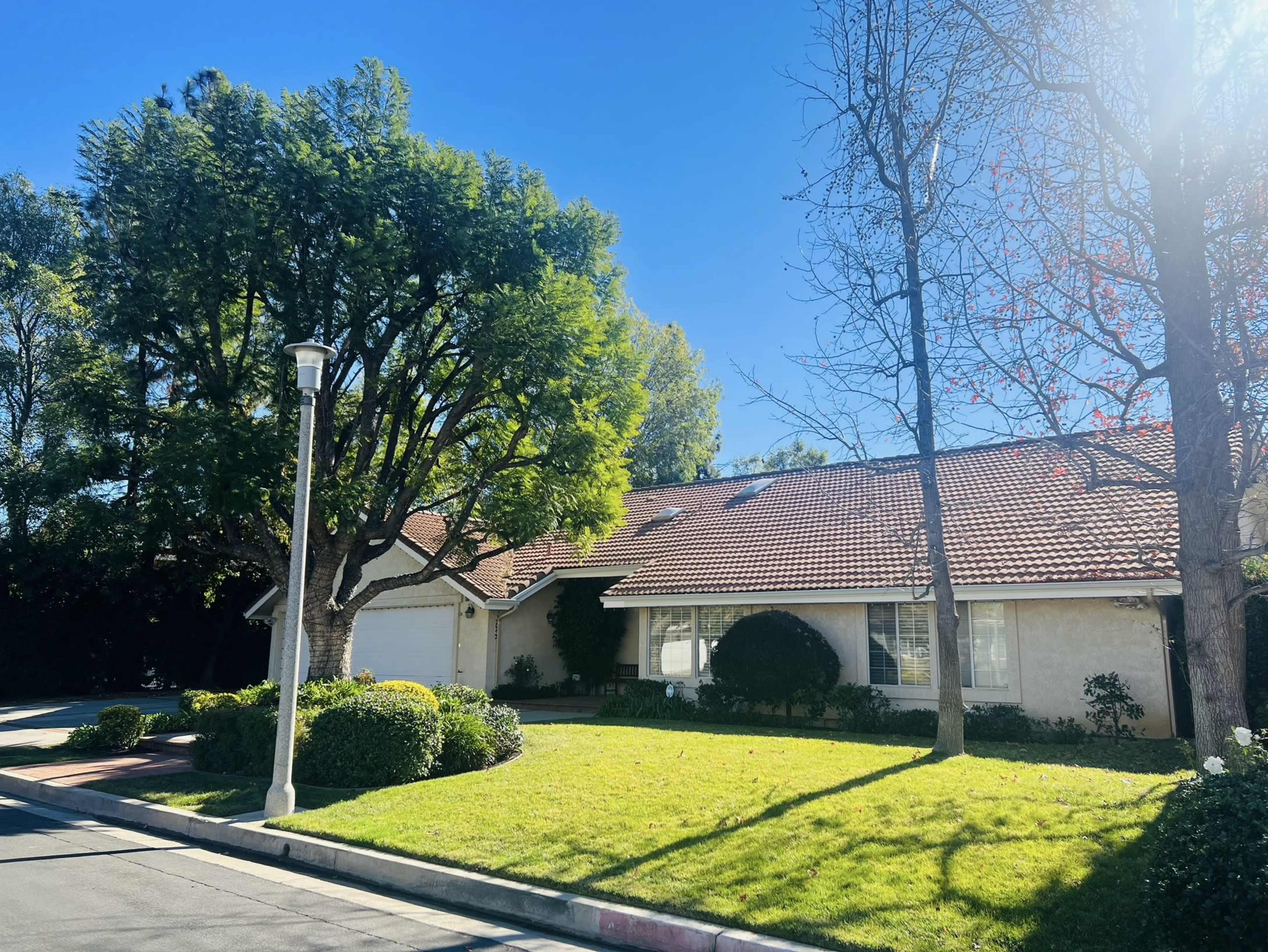 A single-story house with a tiled roof is surrounded by well-maintained grass and shrubs under a clear blue sky.