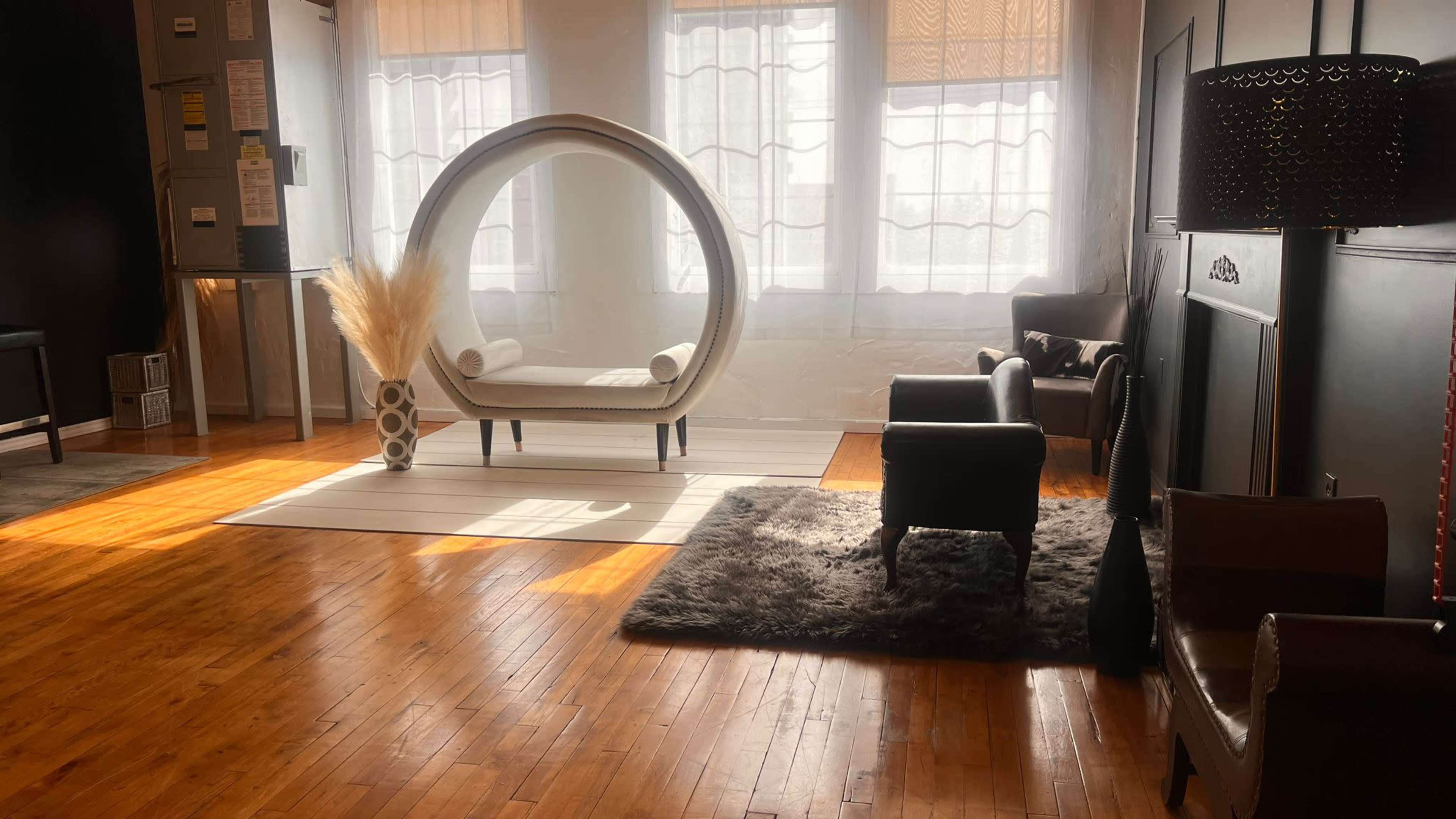 A modern, well-lit room features a circular lounge chair, a plush rug, and various seating options arranged around large windows.