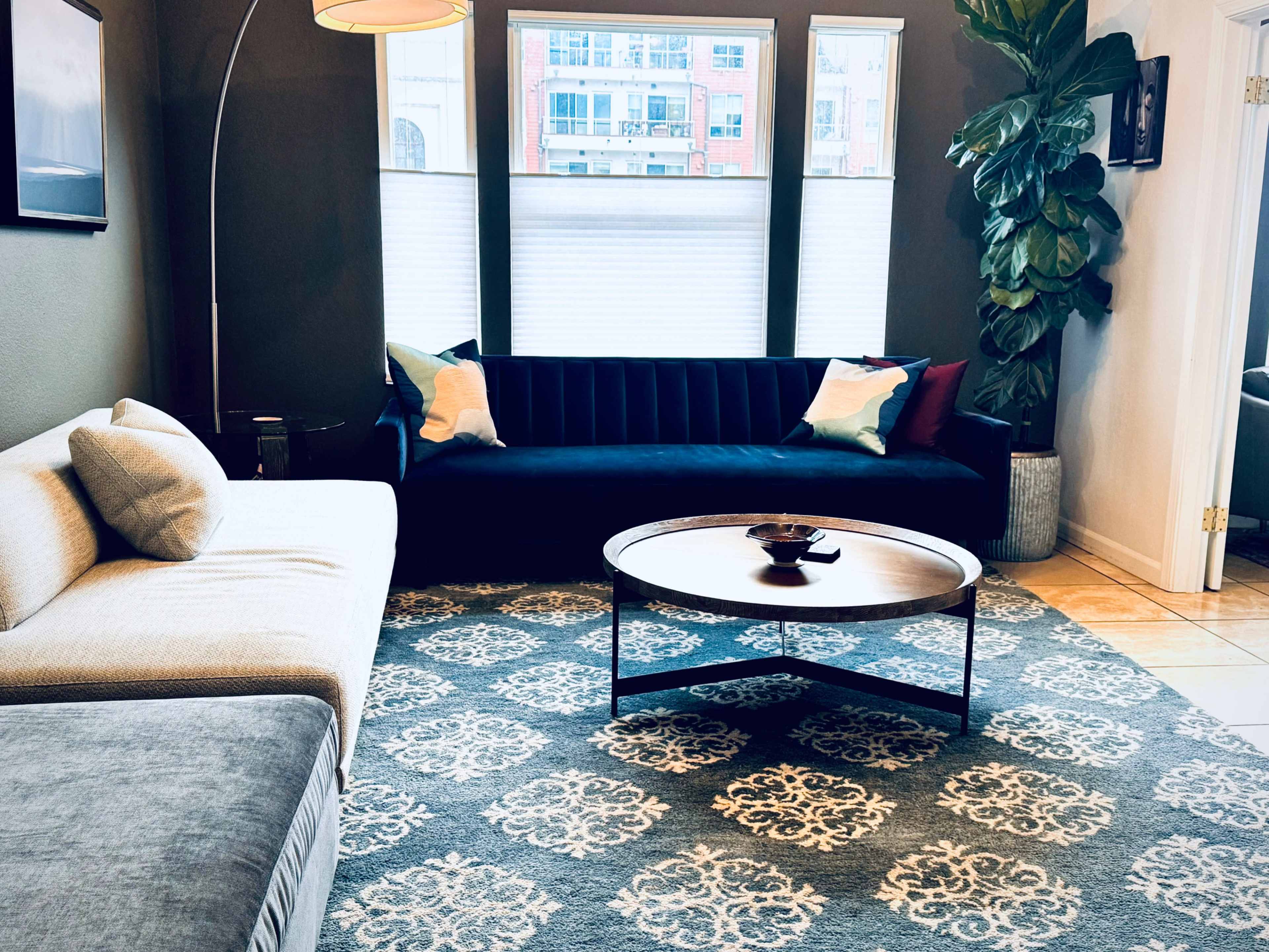 A modern living room with a light-colored sofa, a dark blue couch, a round coffee table, and large windows providing daylight illumination.