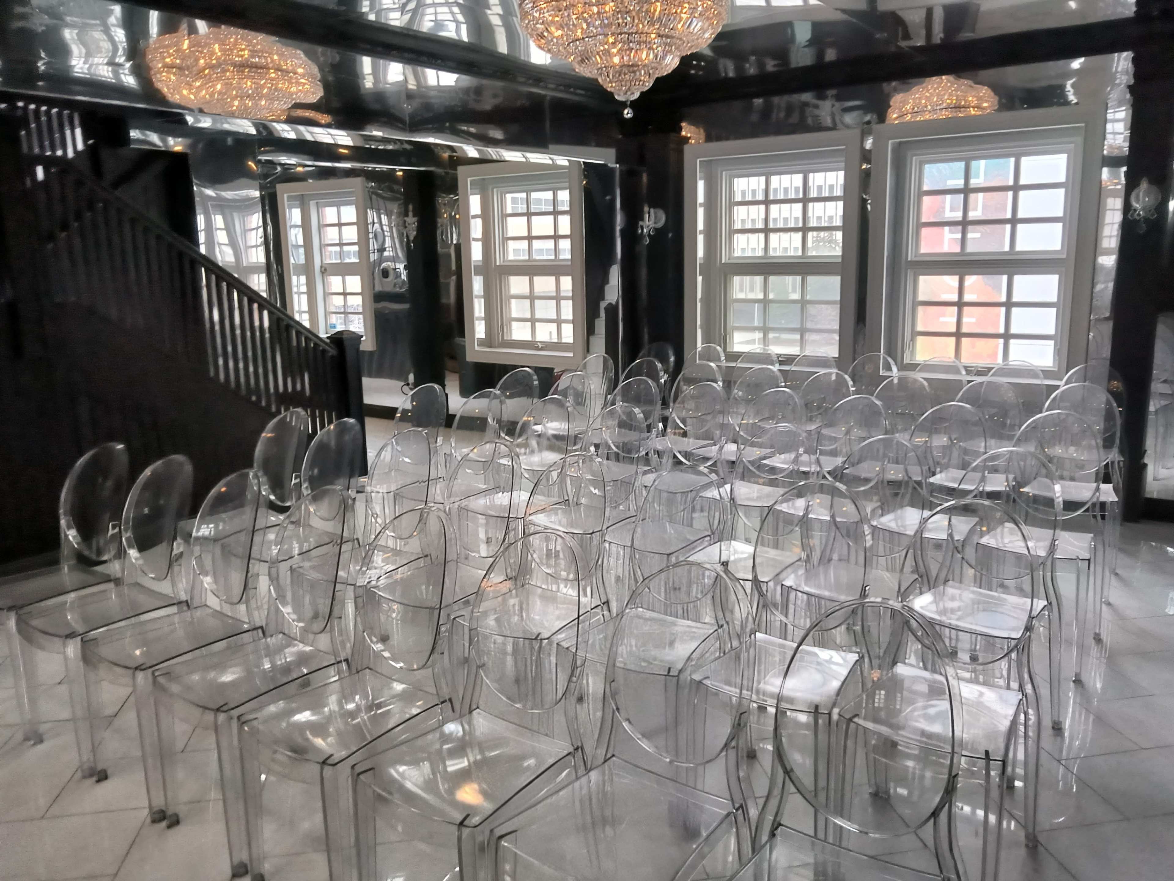 The image shows a room with clear acrylic chairs arranged in rows, under two large crystal chandeliers, with windows visible in the background.