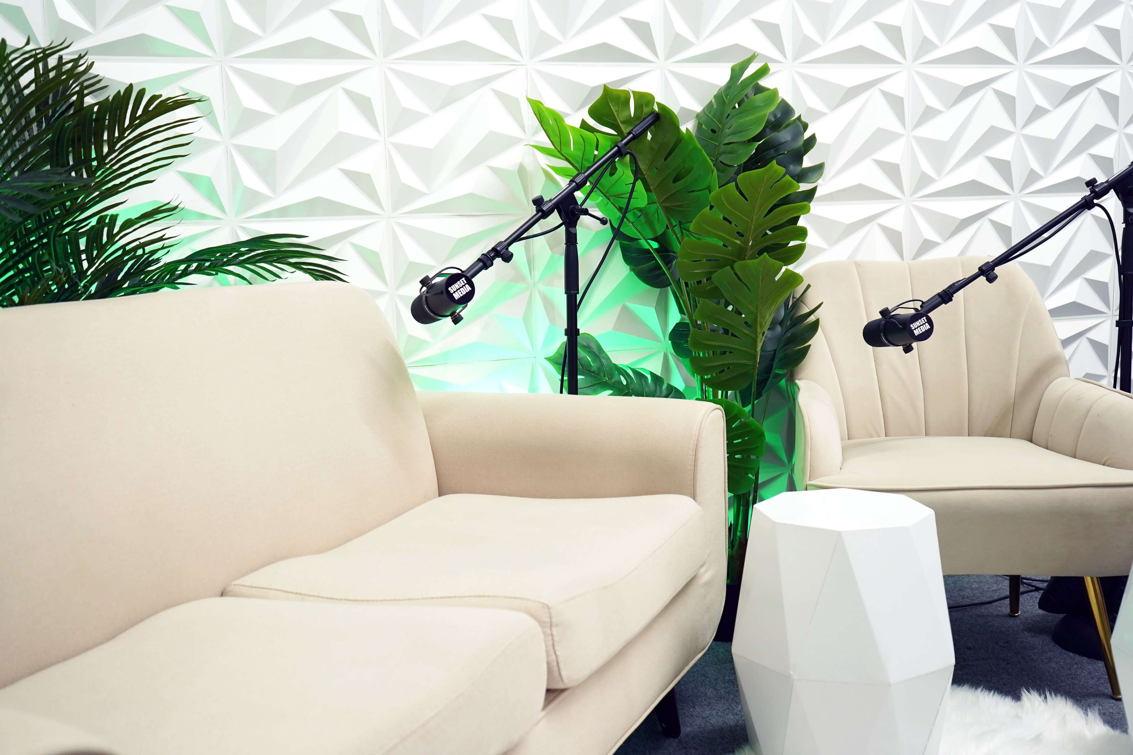 A modern seating area with two light-colored chairs, a geometric white side table, and green plants in front of a textured white wall.
