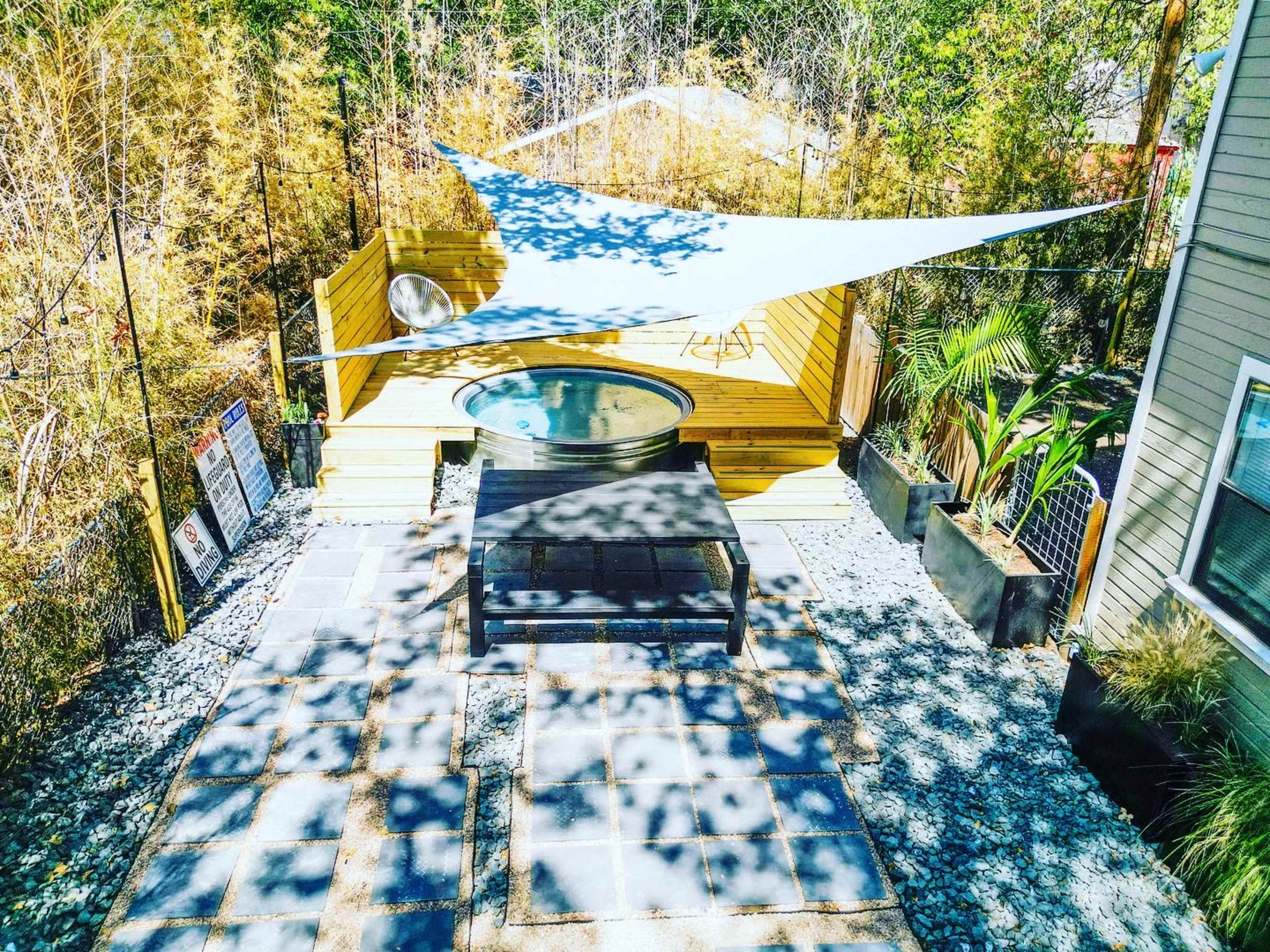 The image shows a wooden deck with a hot tub, a shaded seating area under a sail canopy, and a stone-paved outdoor space surrounded by greenery.