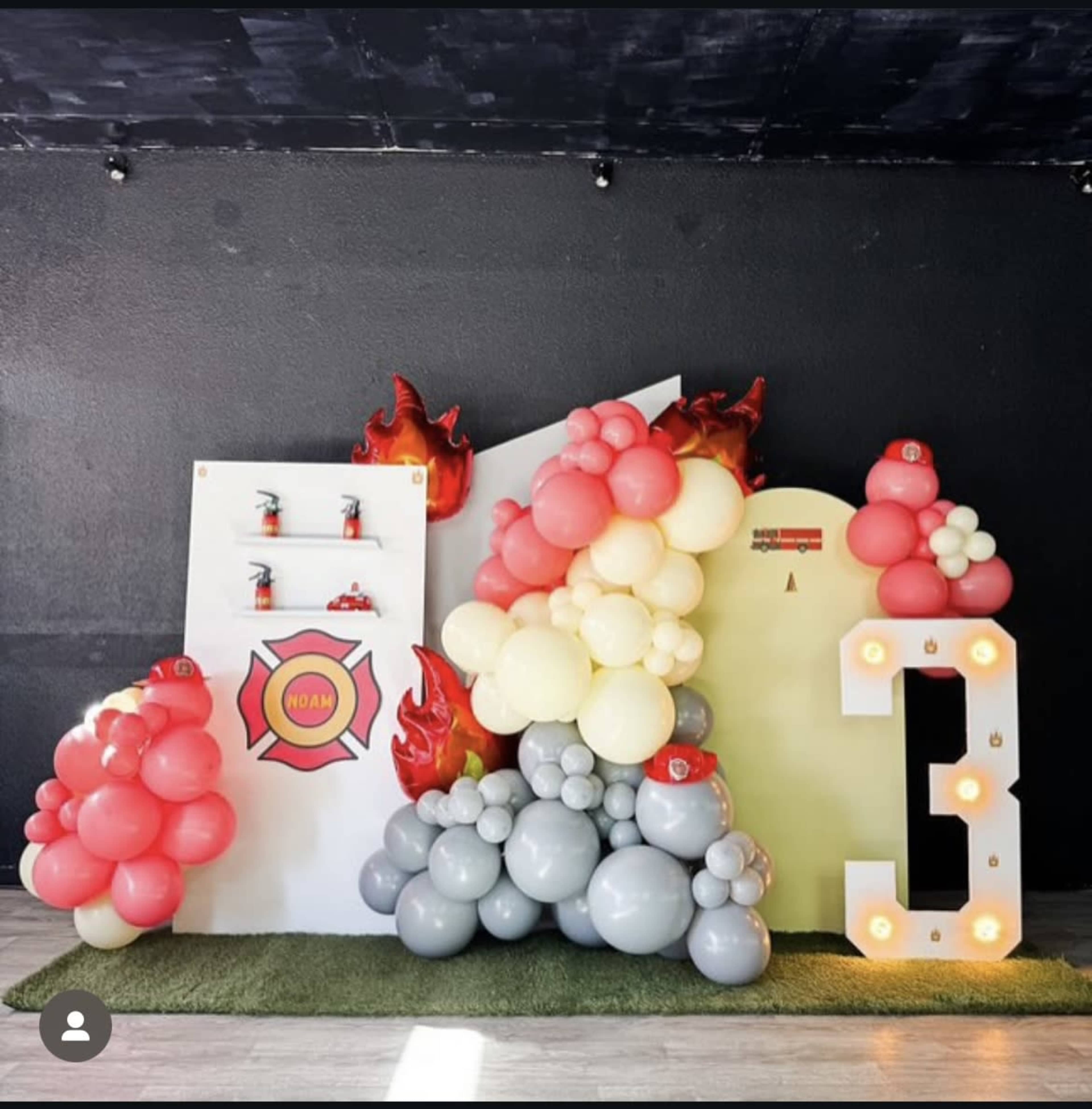 The image shows a decorative setup featuring colorful balloons arranged in clusters with a flame motif, a large number three, and shelves displaying small items.