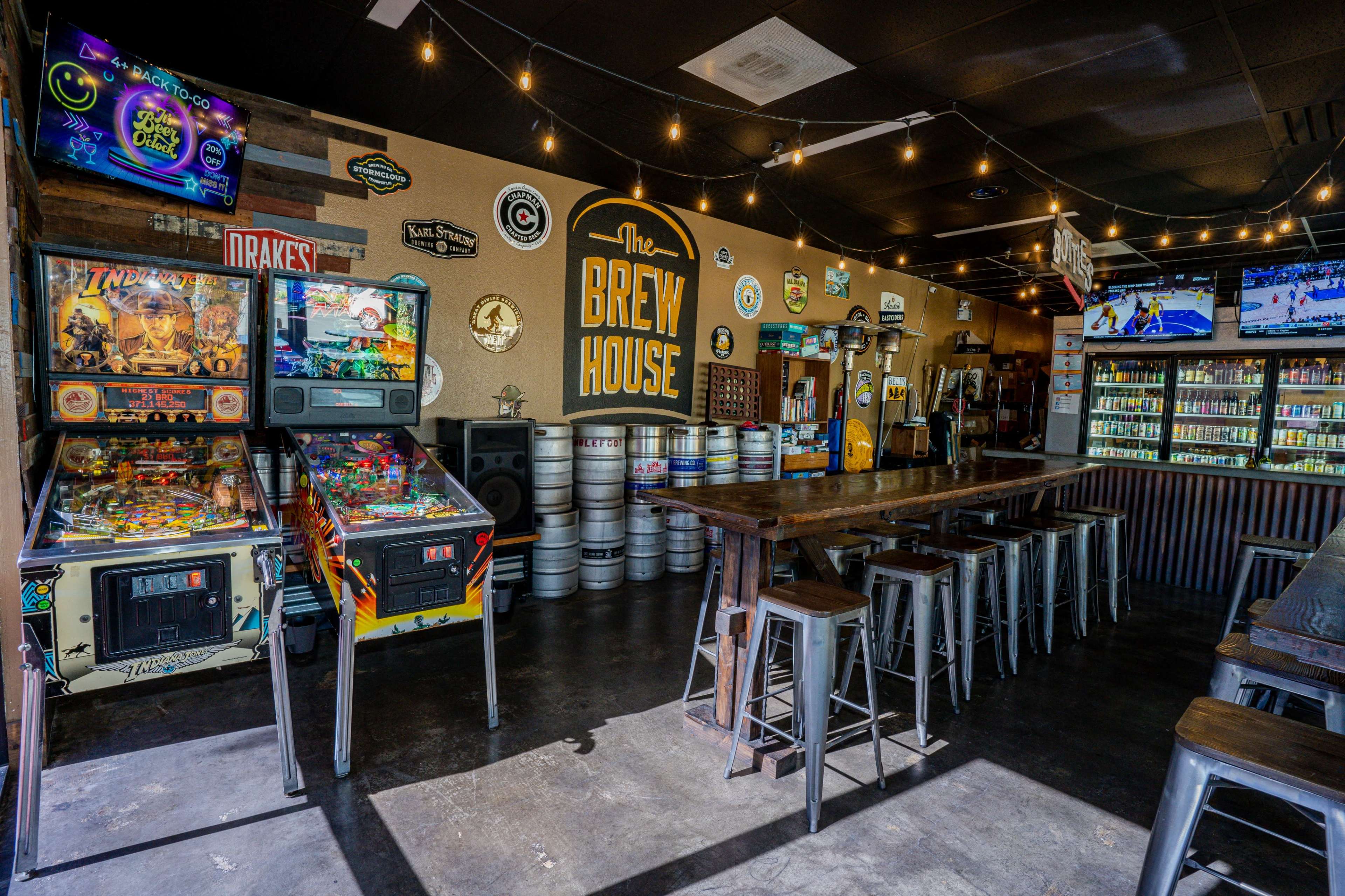 The interior of a brew house features retro pinball machines, a long wooden bar, and various beer taps along the wall.
