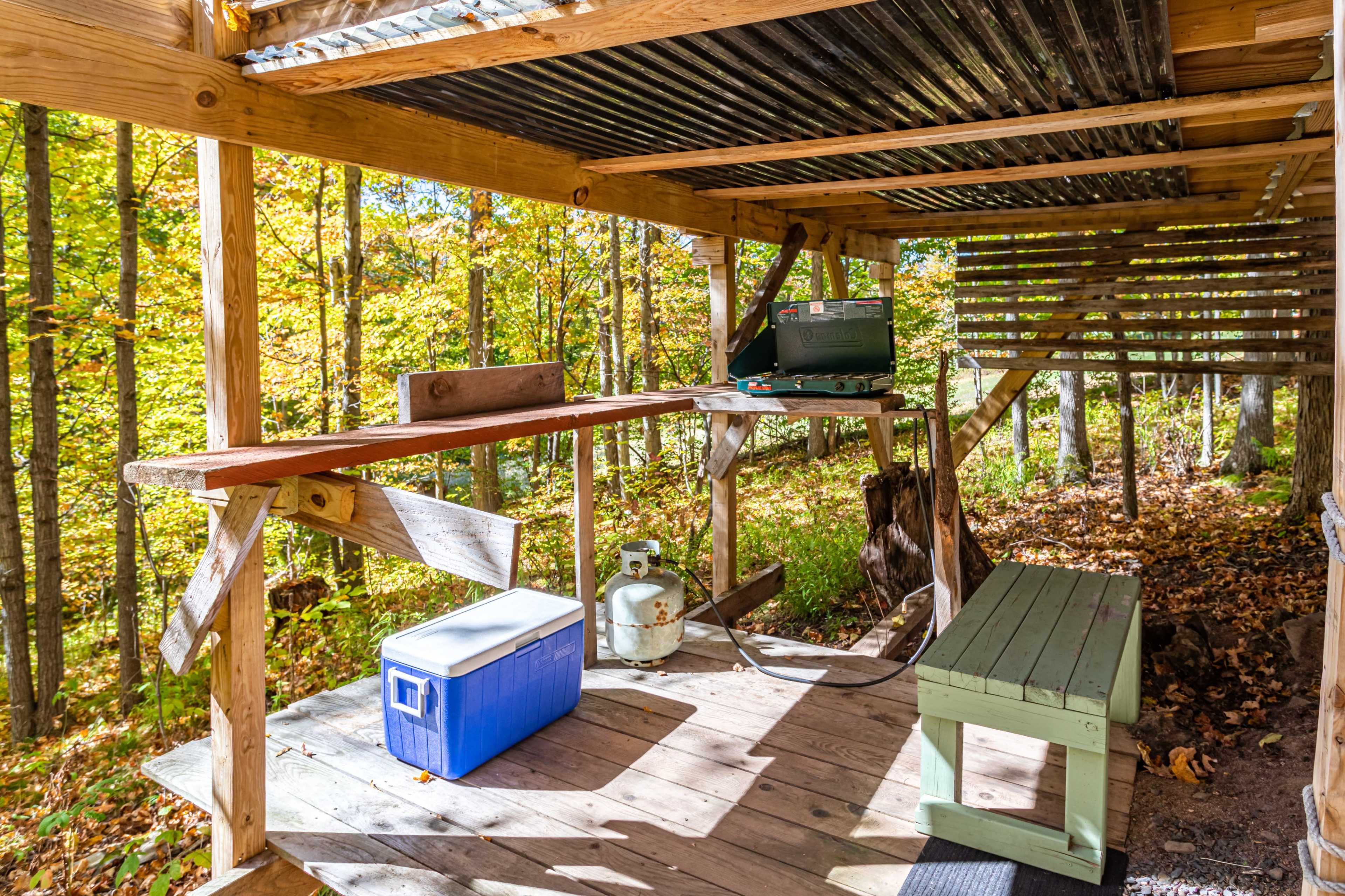 The image shows an open wooden shelter in a forest, furnished with a workbench, a cooler, and a camping stove.