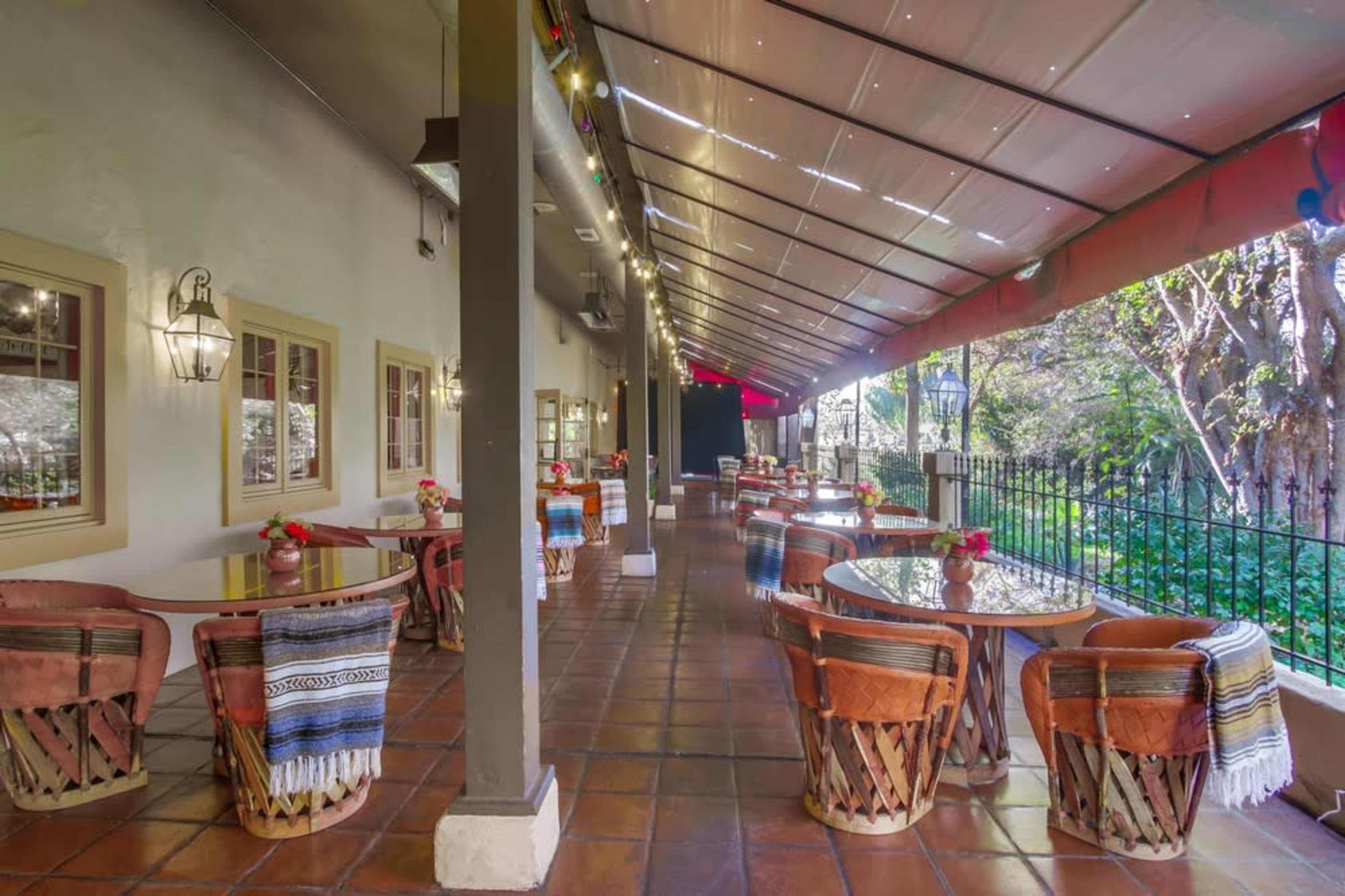 The image shows a covered outdoor dining area with wooden tables and woven chairs, surrounded by greenery and framed by tall windows.