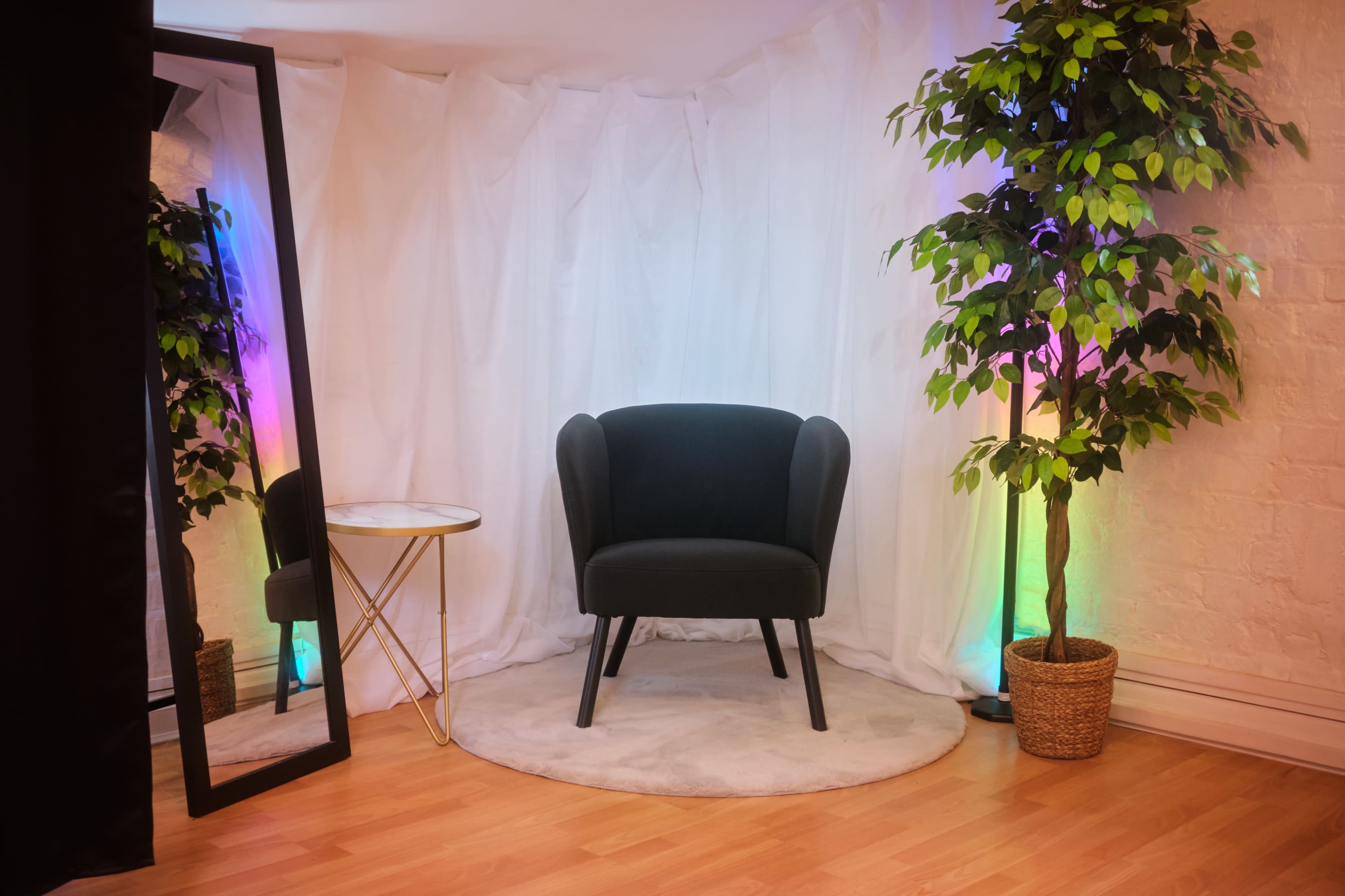 A green upholstered chair is positioned next to a round table and a large mirror, surrounded by a potted plant and draped white fabric with colorful lighting.