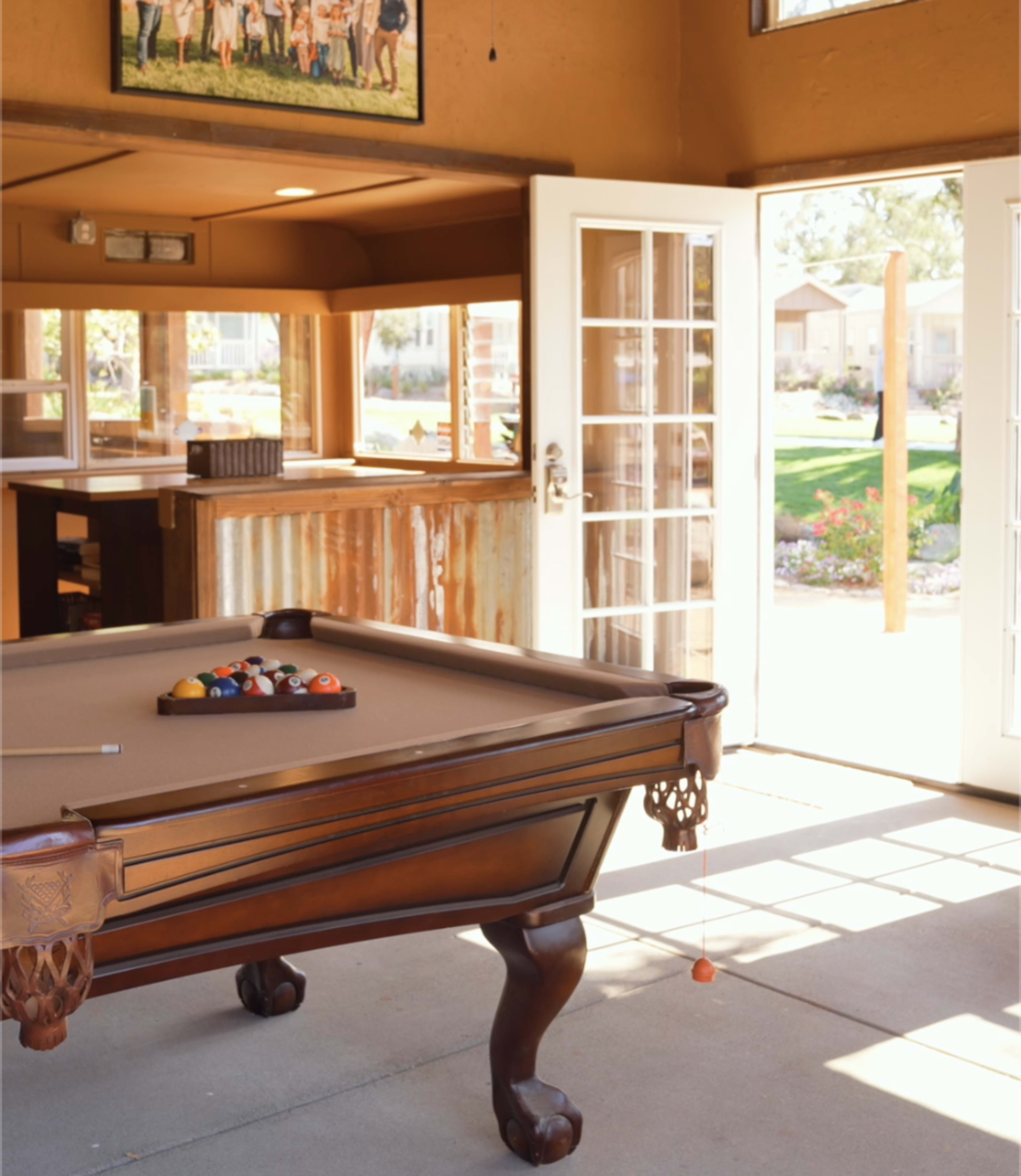 The image shows a pool table in a well-lit room with large windows and an open door leading to an outdoor area.