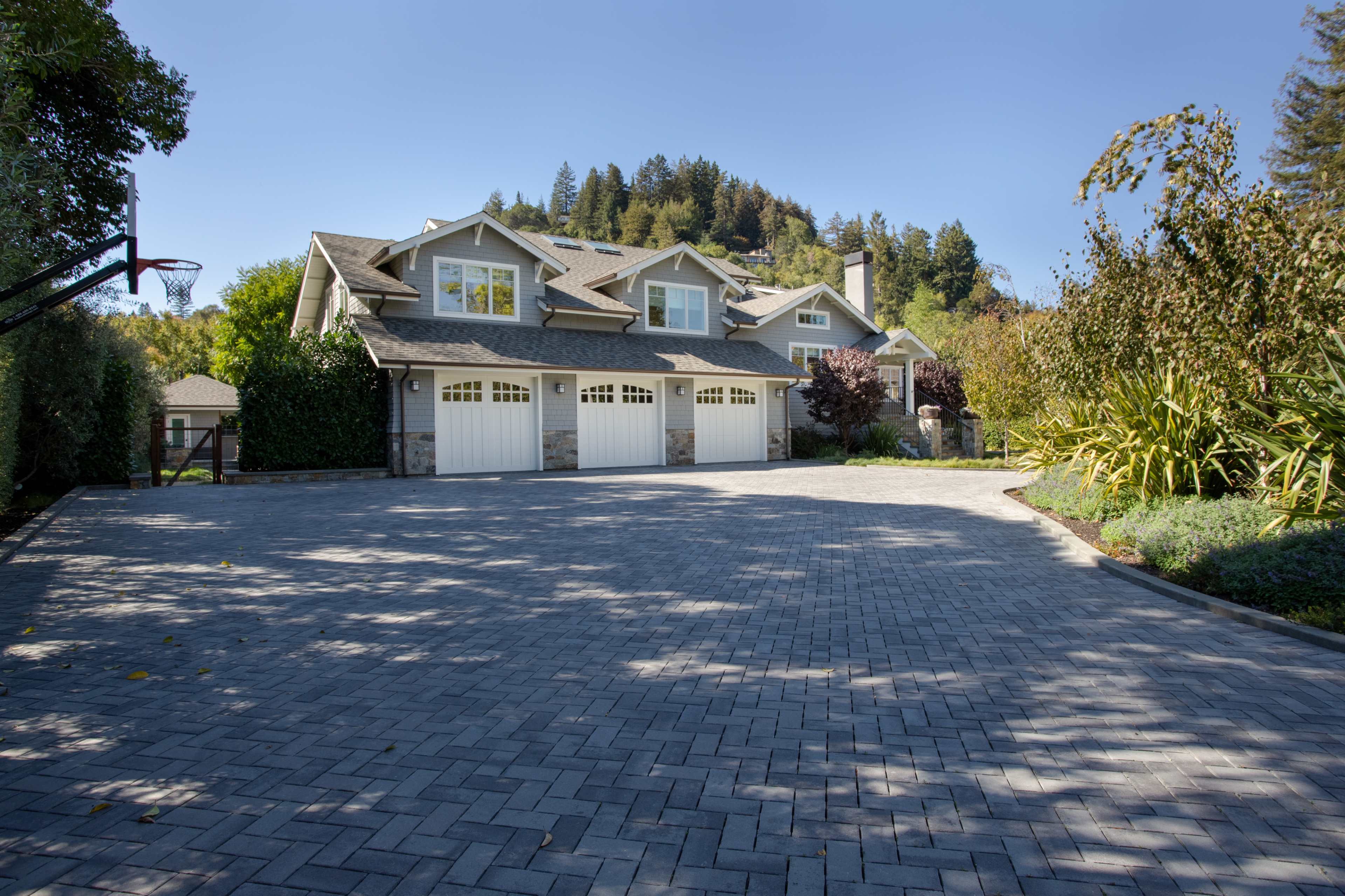 A large, two-story house with a three-car garage sits at the end of a paved driveway surrounded by trees and shrubs.