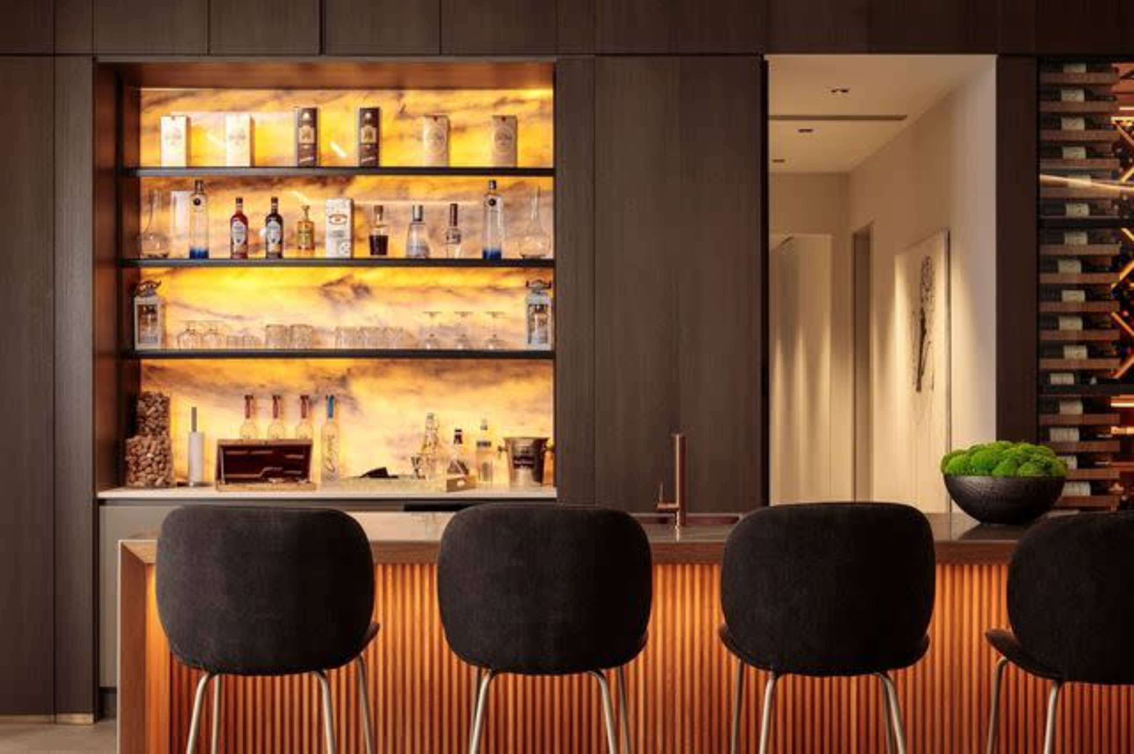 A modern bar features a backlit shelf lined with various bottles and a sleek counter with three upholstered stools.