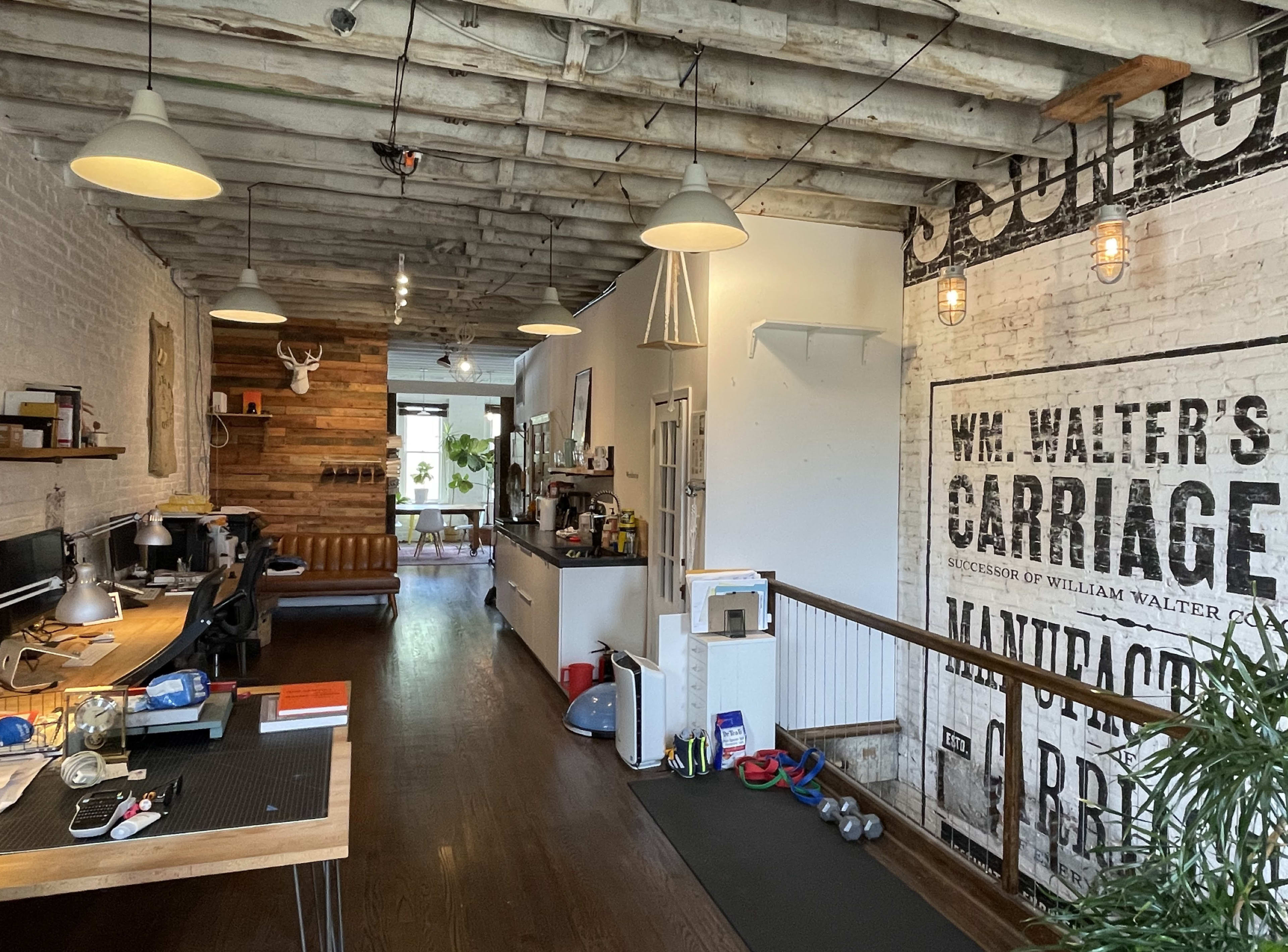 The image shows an open office space with wooden beams, a mix of modern and rustic decor, desks with computers, and exercise equipment in the corner.