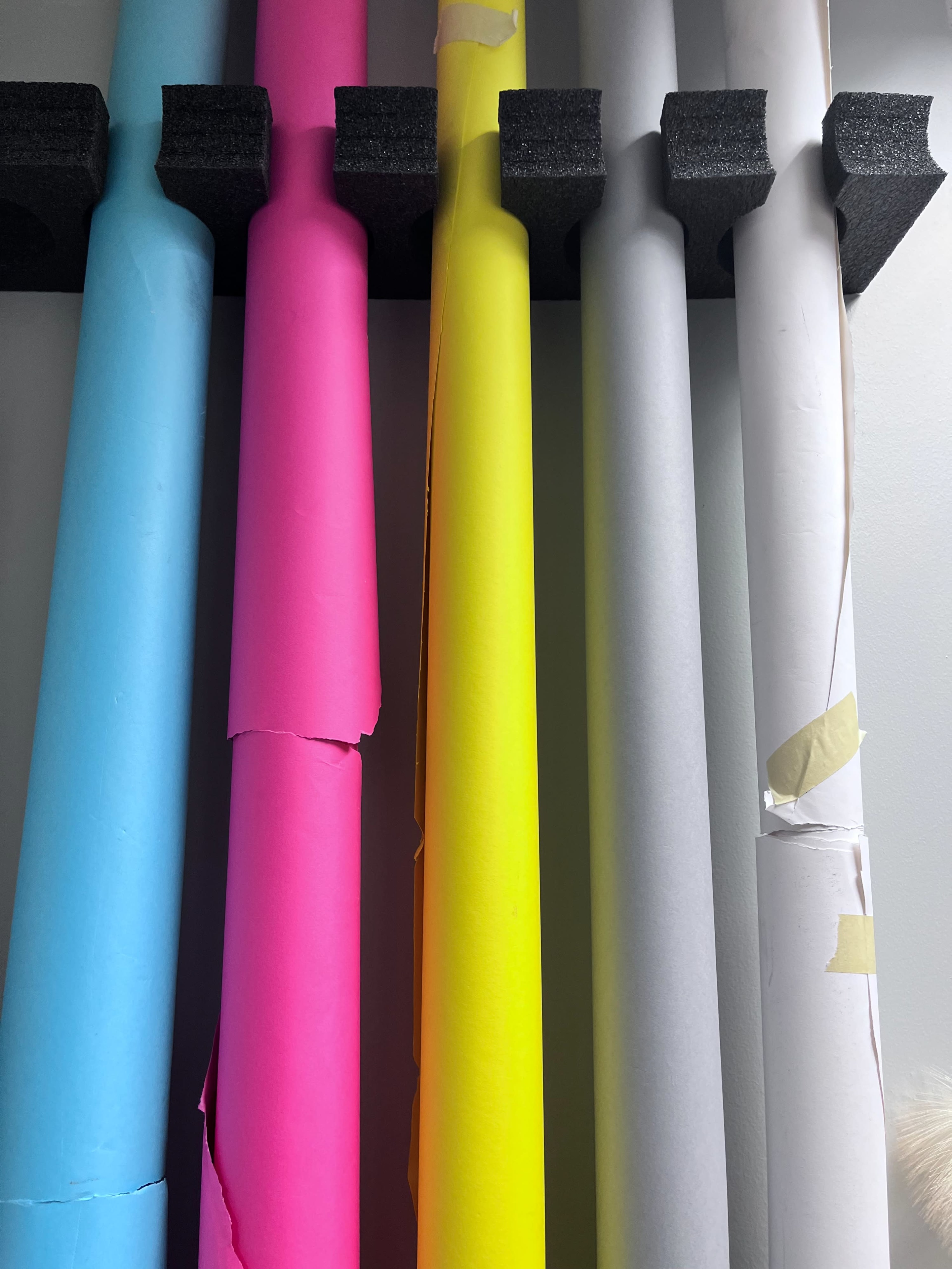 A row of rolled paper and backdrop materials in various colors, including blue, pink, yellow, gray, and white, resting on a foam holder against a wall.