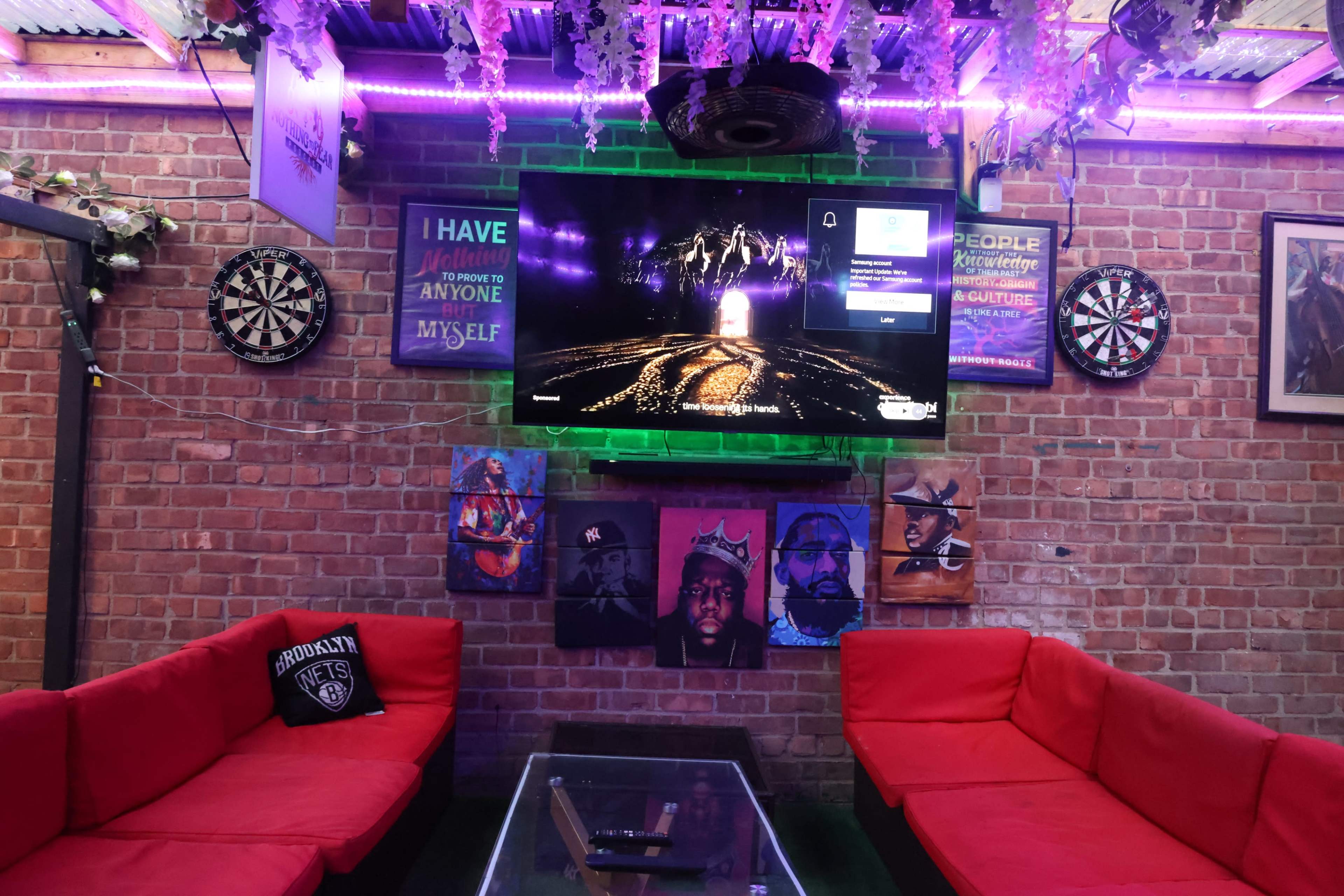 The image shows a room with brick walls, a large television mounted on the wall, and red couches arranged around a glass coffee table, with dartboards and framed artwork visible.