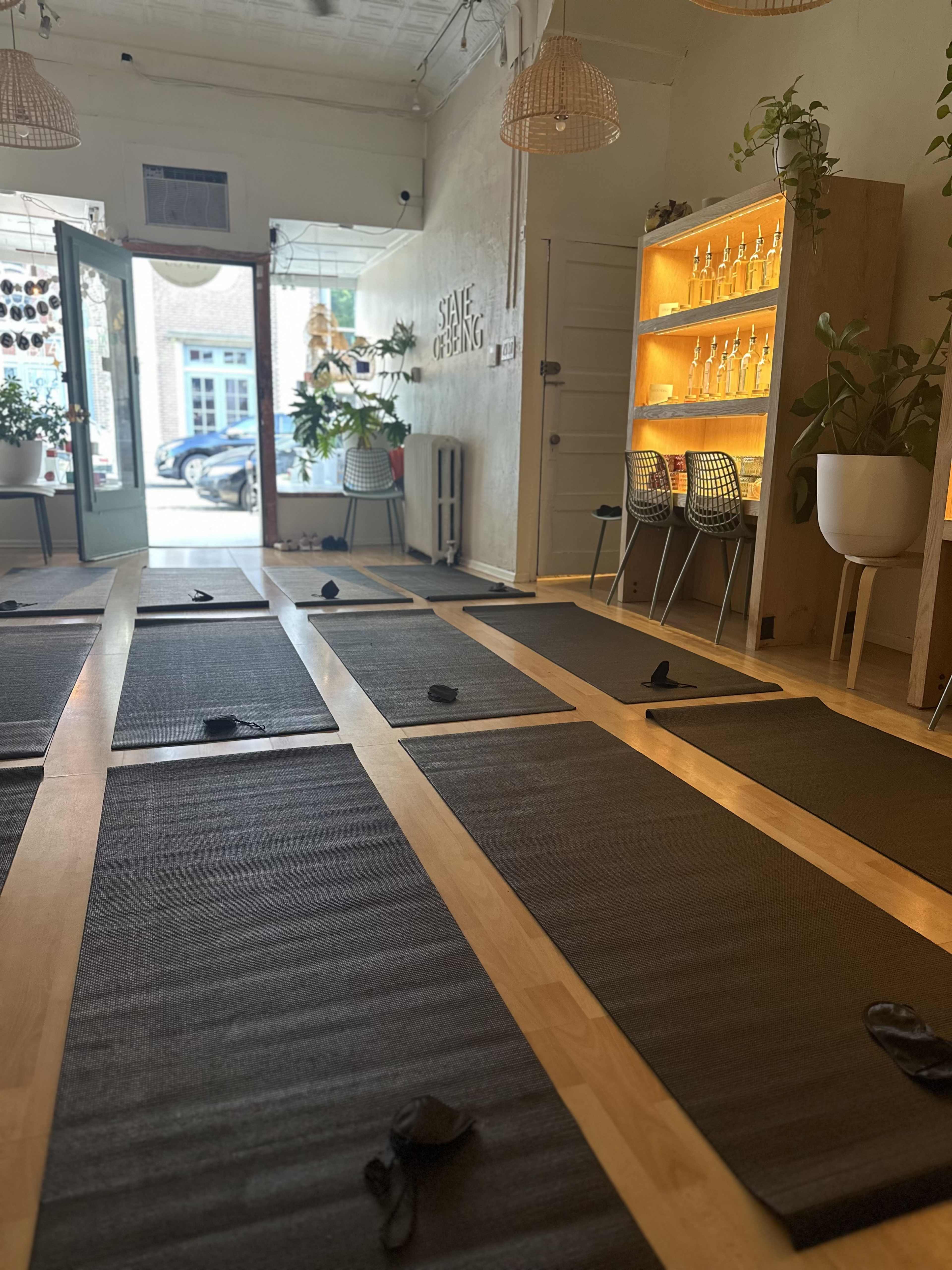 The image shows a yoga studio with several mats arranged on a wooden floor, and a reception area featuring plants and a backlit display shelf.