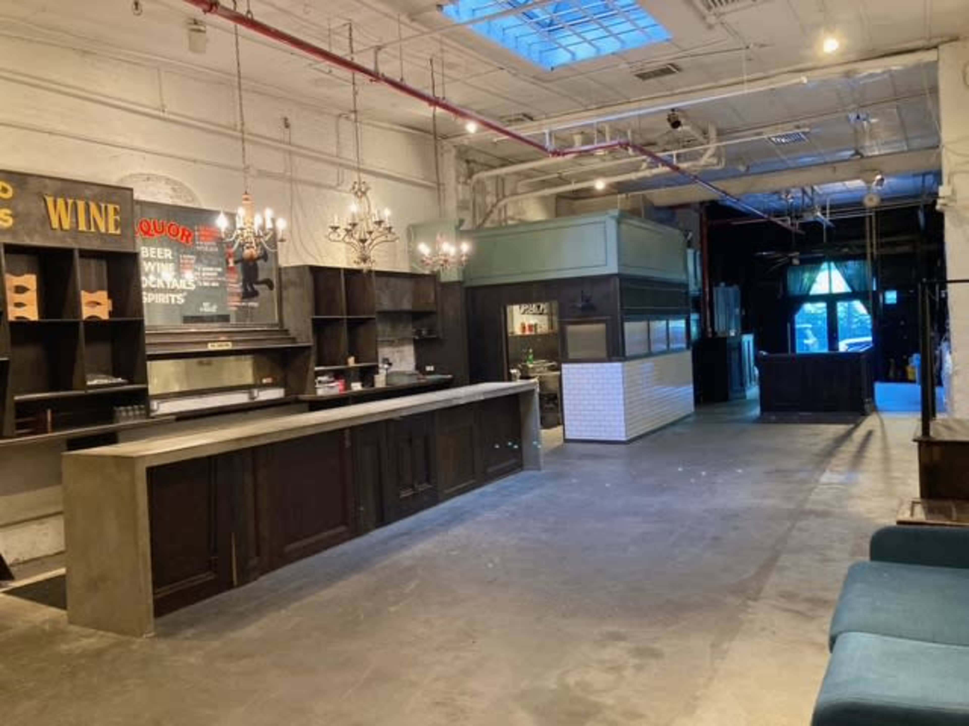 The image shows an empty interior space with a bar area featuring a long countertop, shelves, and chandeliers, along with a section that appears to be a kitchen or serving area, all set in a well-lit, industrial-style environment.