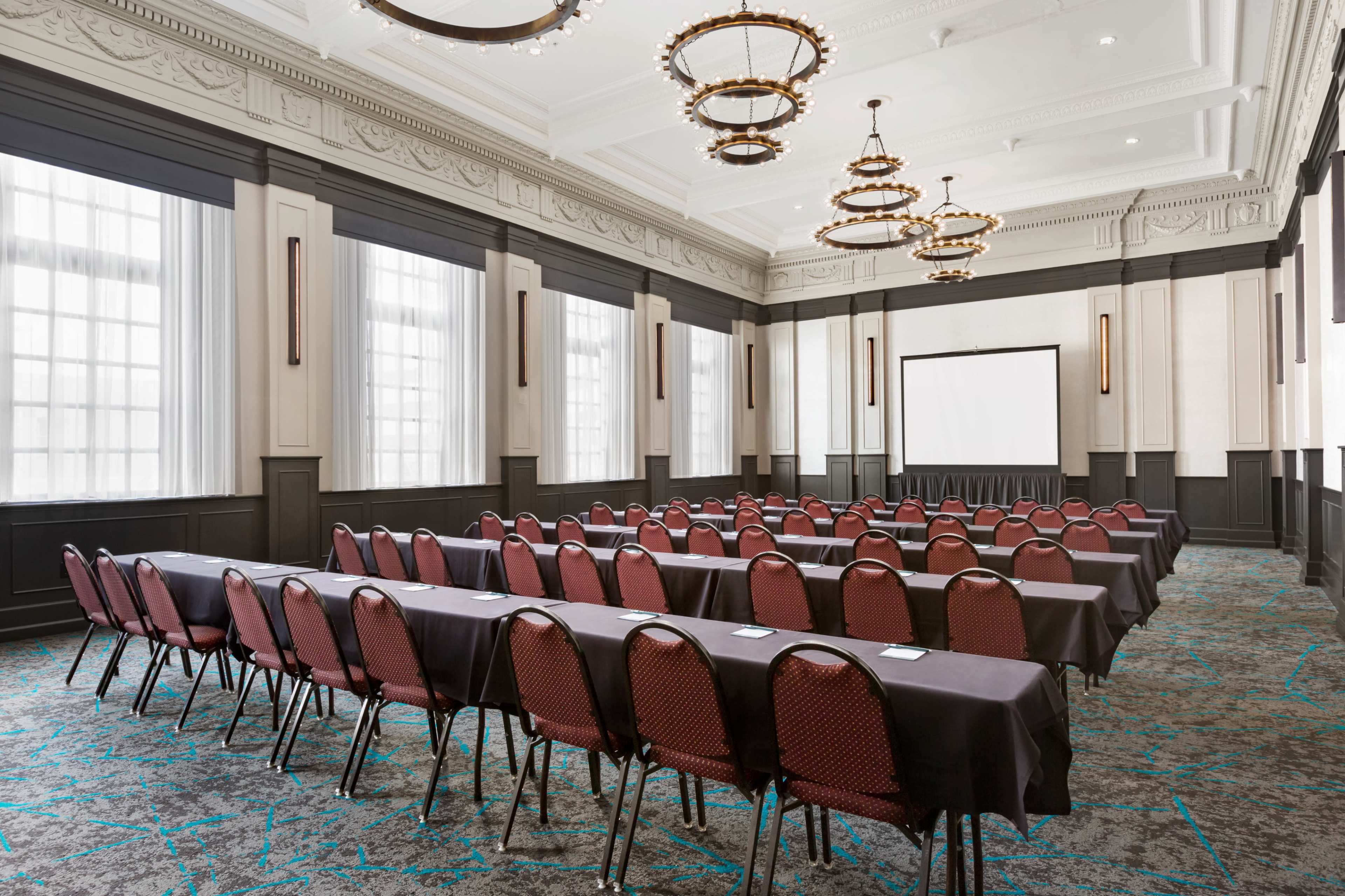 The image shows a spacious conference room arranged with rows of tables and chairs, featuring a large screen at the front for presentations.