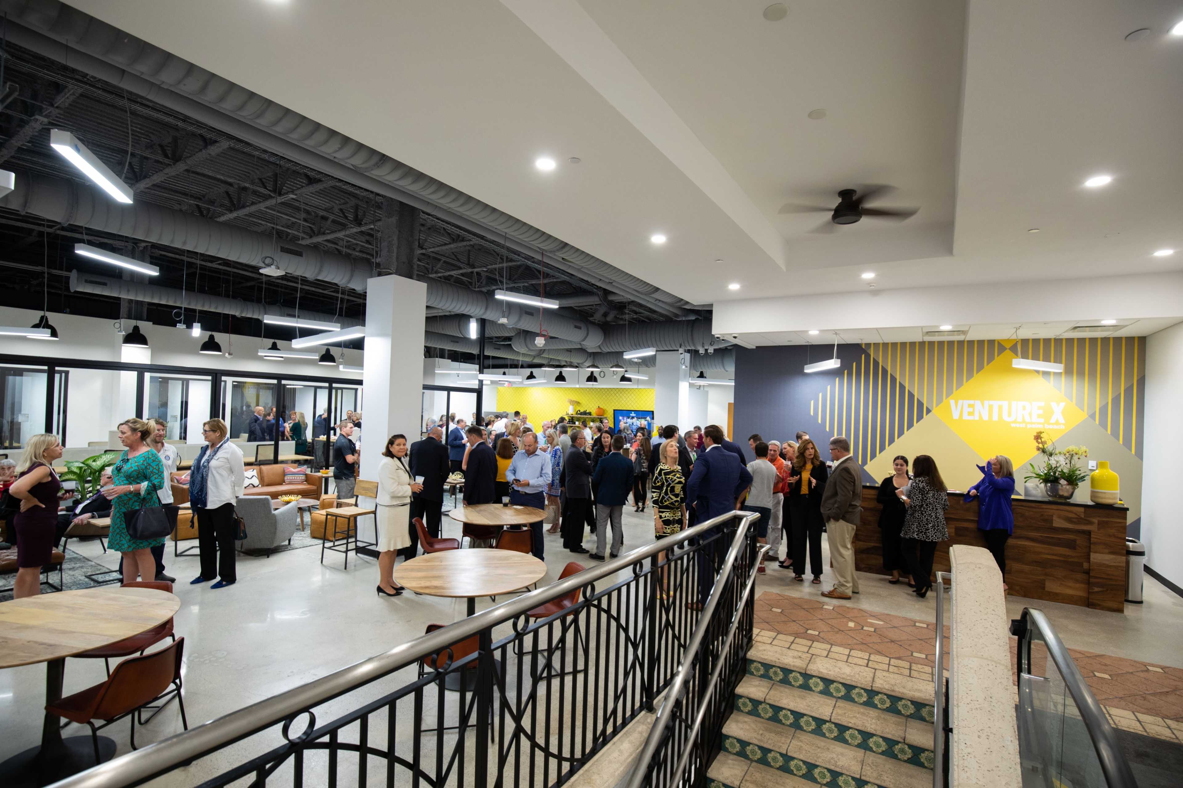 A large group of people network and socialize in a modern coworking space with various seating areas and a vibrant yellow accent wall.