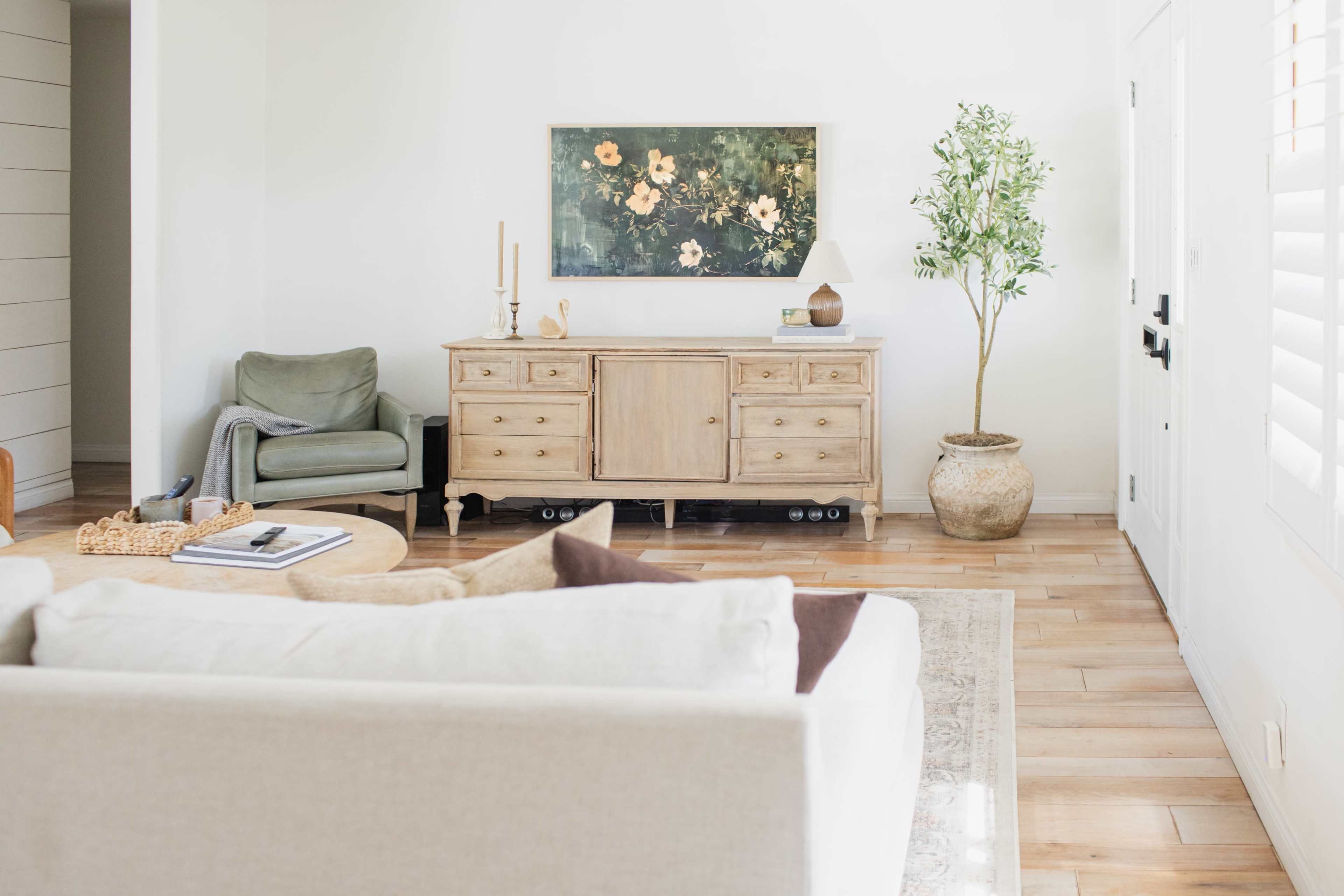The image shows a bright, minimalist living room with a wooden sideboard, a green armchair, a round coffee table, and a potted plant.