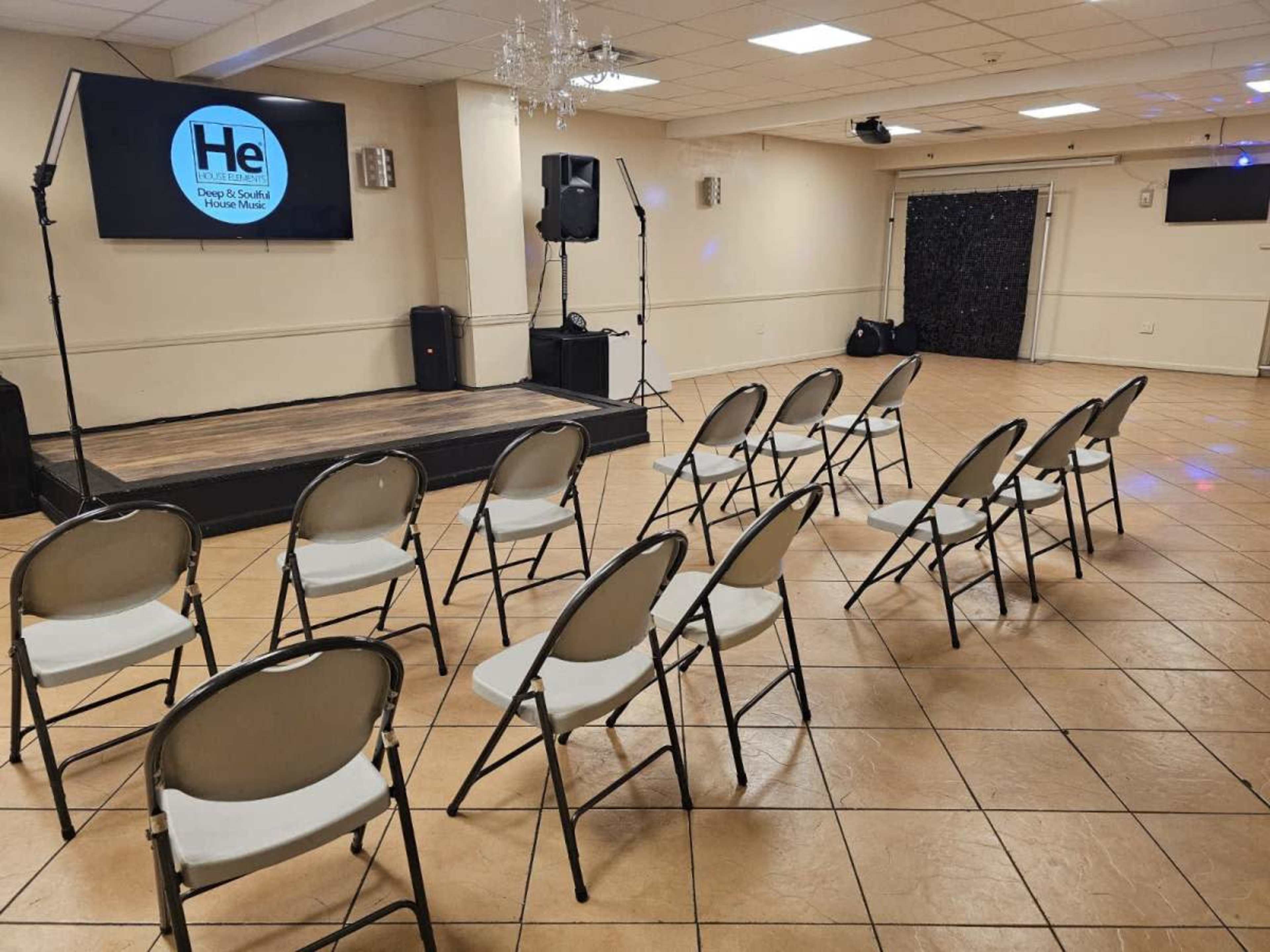 A meeting room with a raised stage, a large screen displaying a logo, and several rows of empty folding chairs arranged in front.