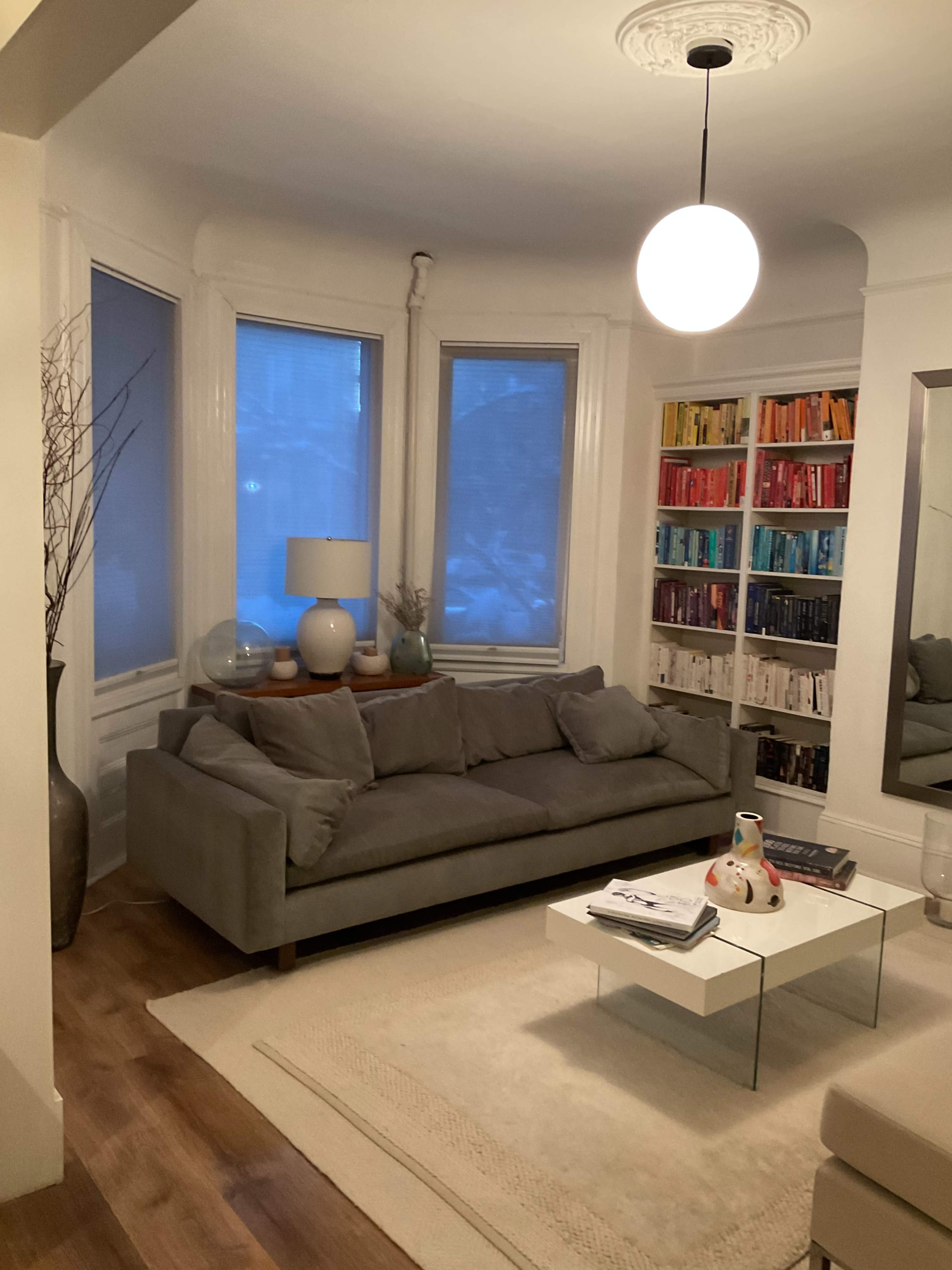 The image depicts a cozy living room featuring a gray sofa, a glass coffee table, and a bookshelf filled with colorful books, all illuminated by a hanging pendant light.