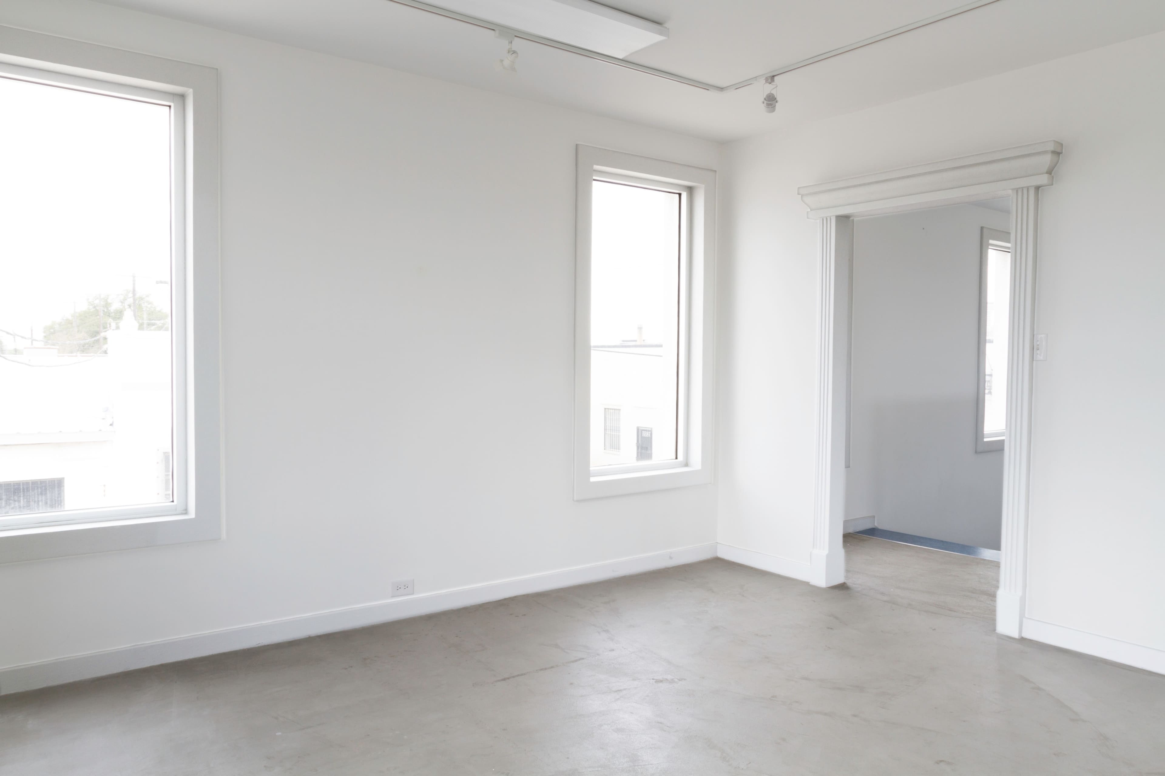 The image shows a minimalist, empty room with large windows and a doorway leading to another room.