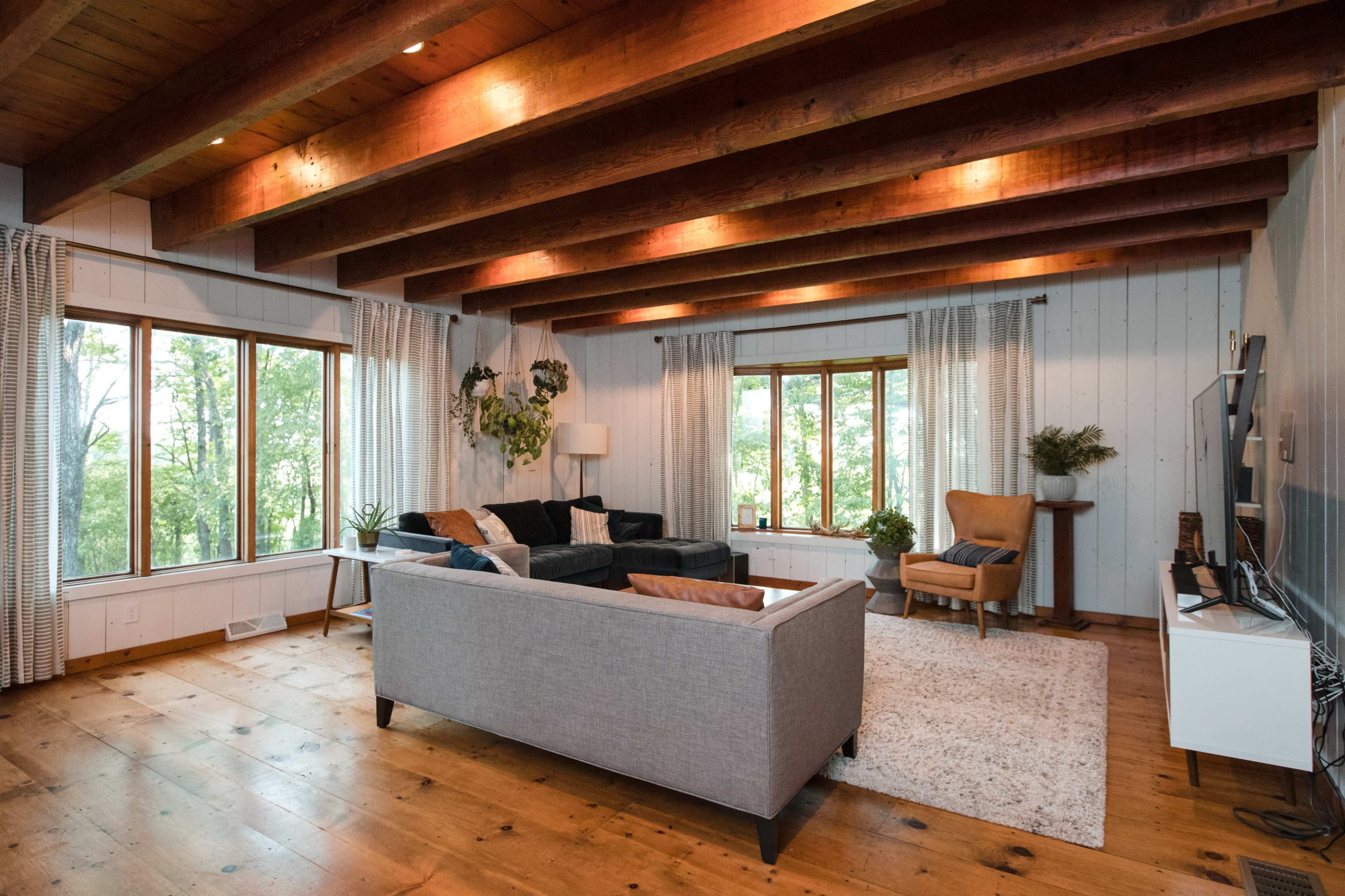A modern living room features large windows with views of greenery, wooden beams on the ceiling, and a mix of furniture including a couch, armchair, and a TV stand.