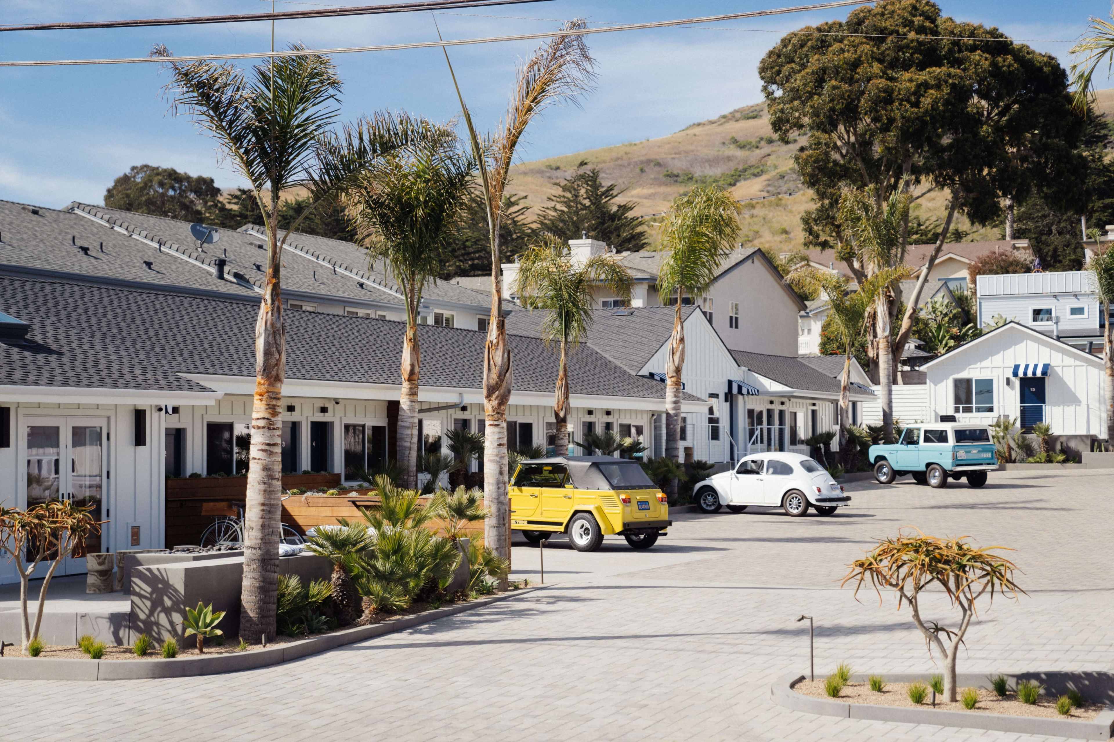 A row of single-story buildings with palm trees and parked vintage cars is situated in a paved lot against a backdrop of rolling hills.