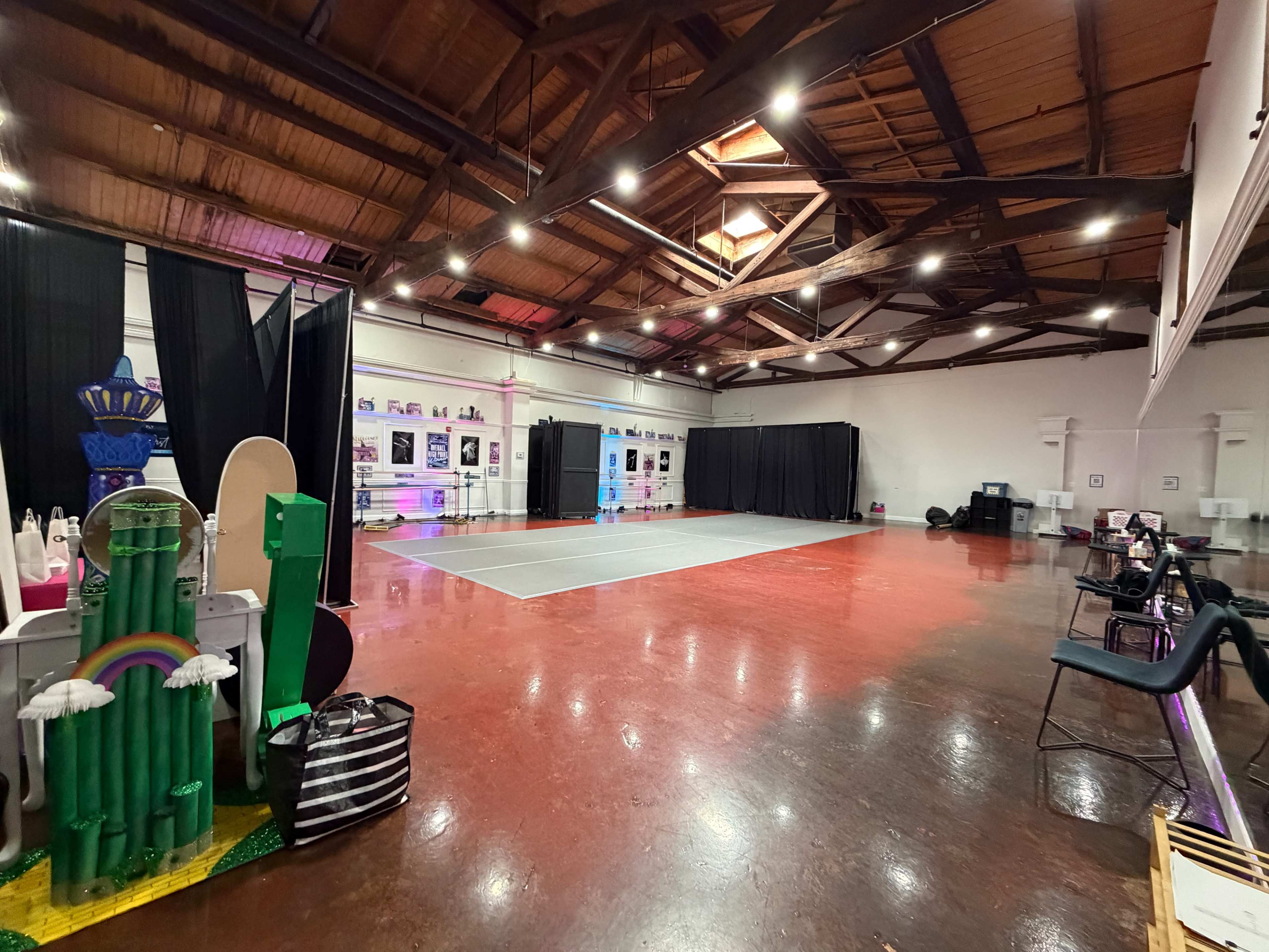 The image depicts a spacious indoor hall with polished floors, featuring a gray dance area in the center and various decorations along the side, including colorful structures and black curtains.