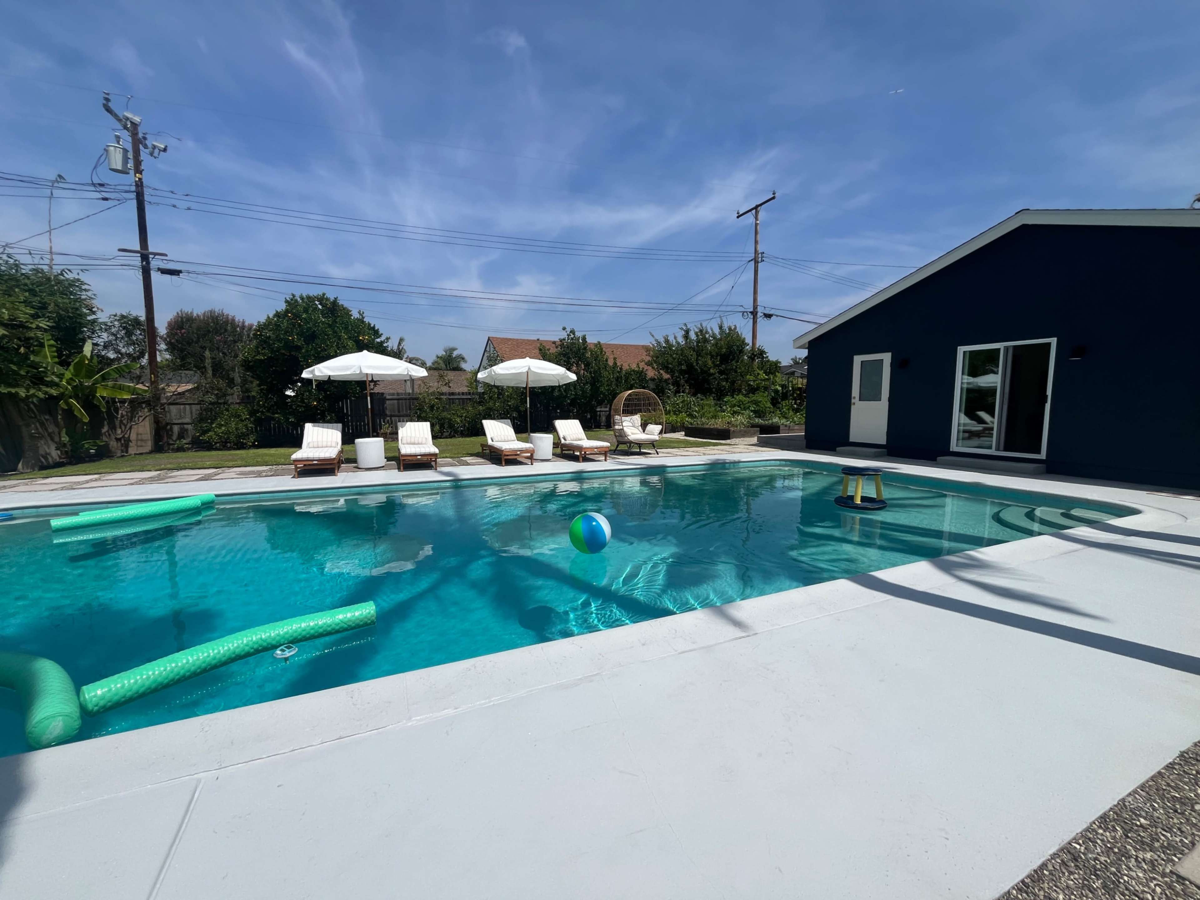 The image shows a spacious swimming pool surrounded by lounge chairs and a house with large windows on a sunny day.