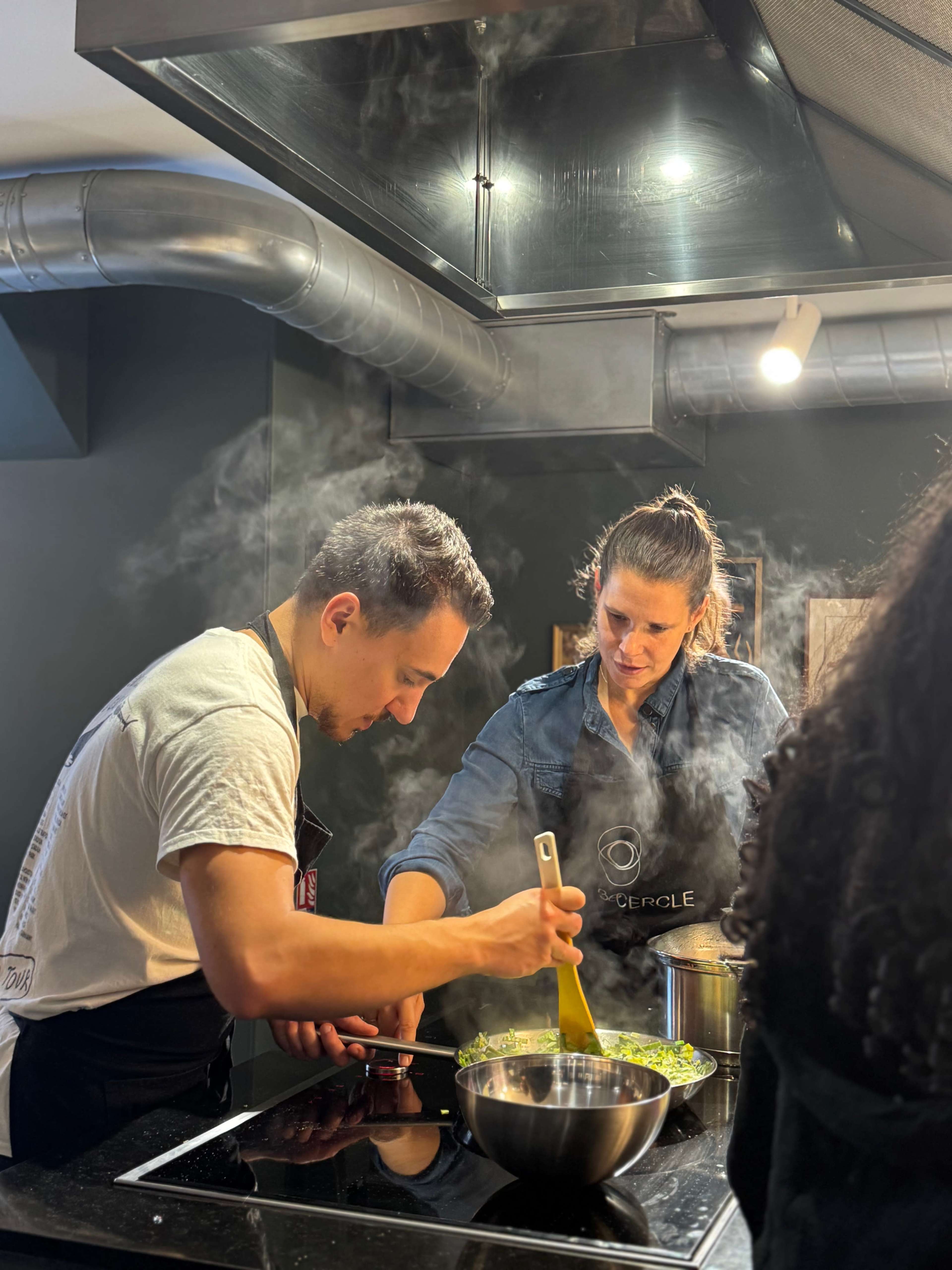 Two people are cooking in a modern kitchen, working together over a bowl of chopped vegetables while steam rises from pots on the stove.