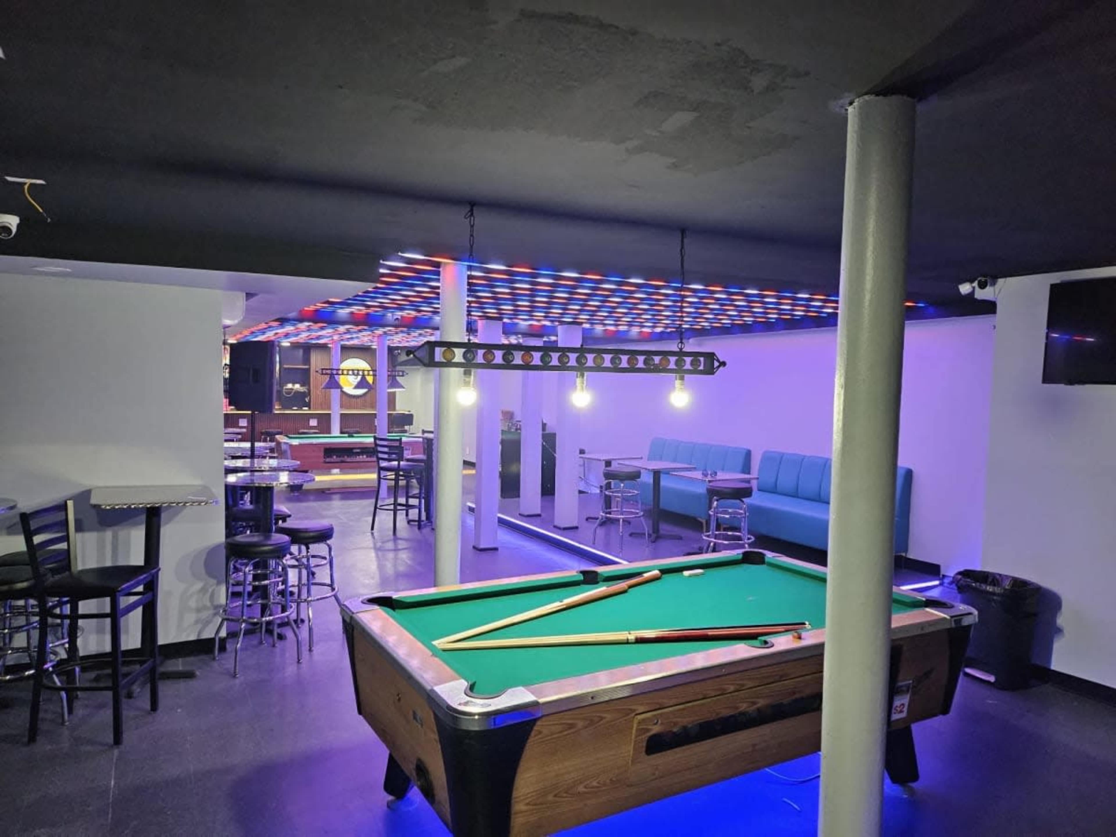 The image shows a dimly lit pool hall featuring a green felt pool table, bar stools, and a seating area under colorful LED lights.