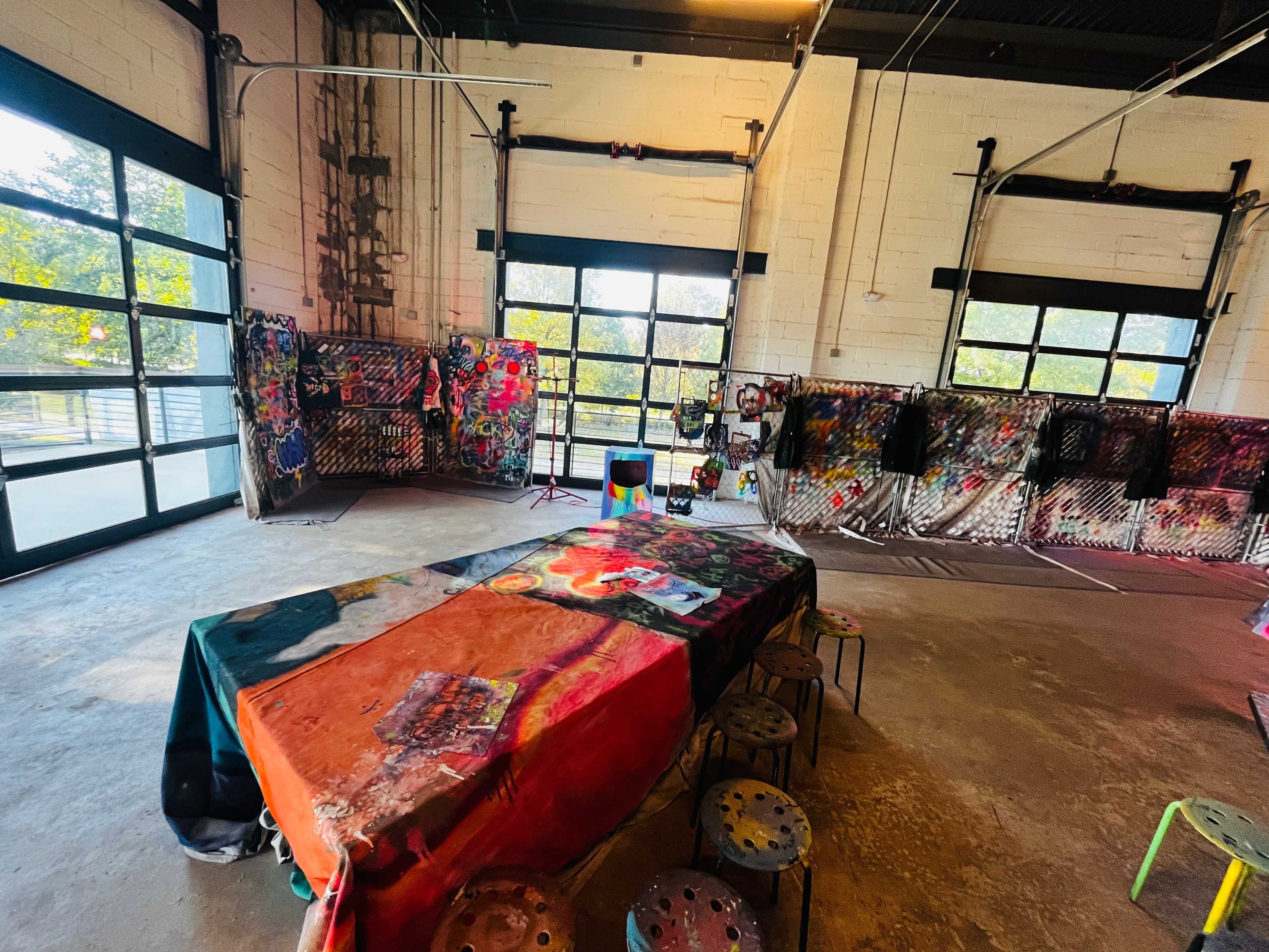 An art studio with a colorful table covered in artworks and walls adorned with various paintings and mixed media pieces.