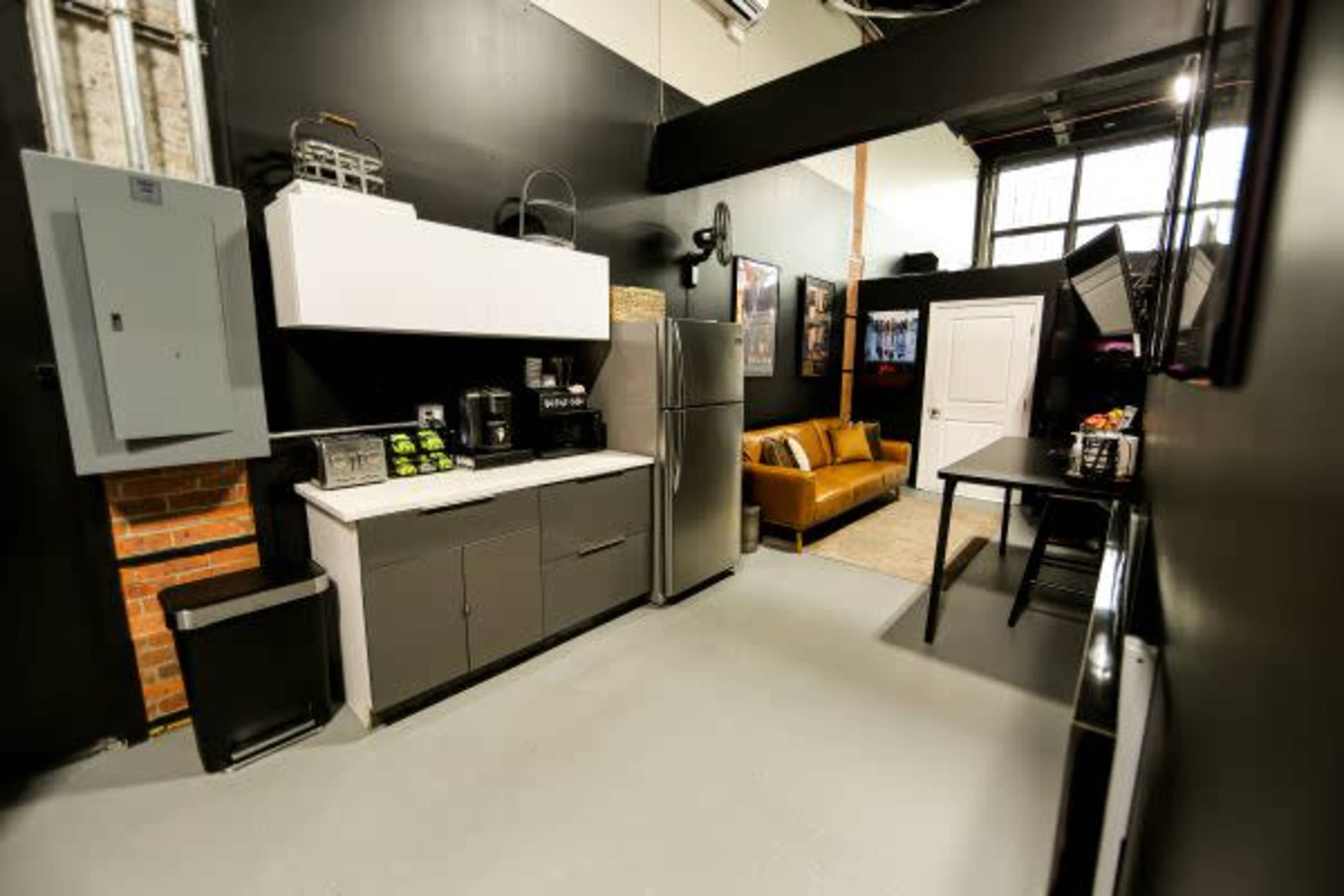 The image shows a modern kitchen space with a refrigerator, coffee maker, and a countertop, adjacent to a living area featuring a brown couch and a small dining table.