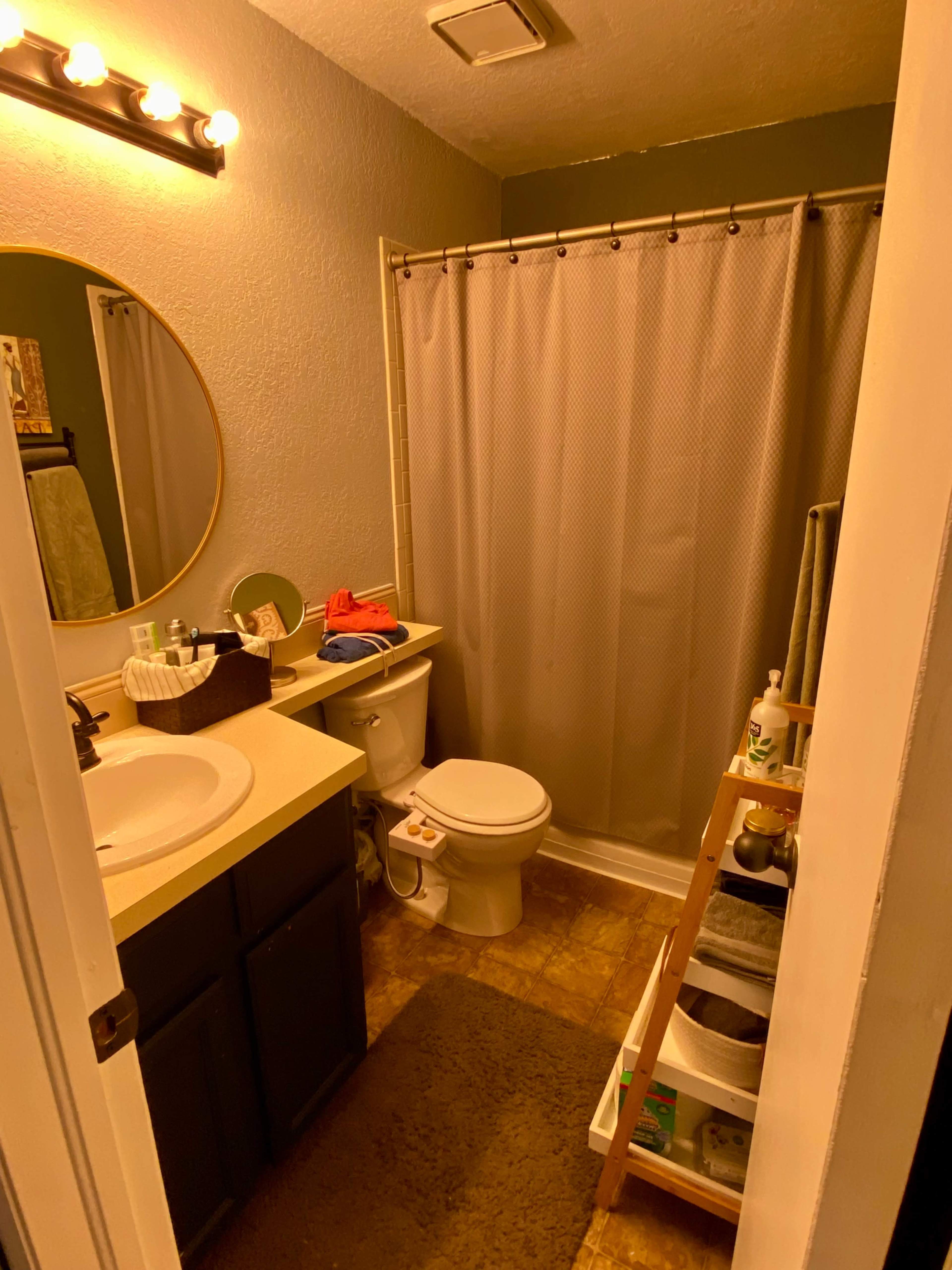 A small bathroom features a circular mirror above a sink, a shower with a curtain, a toilet, and storage shelves with various toiletries.