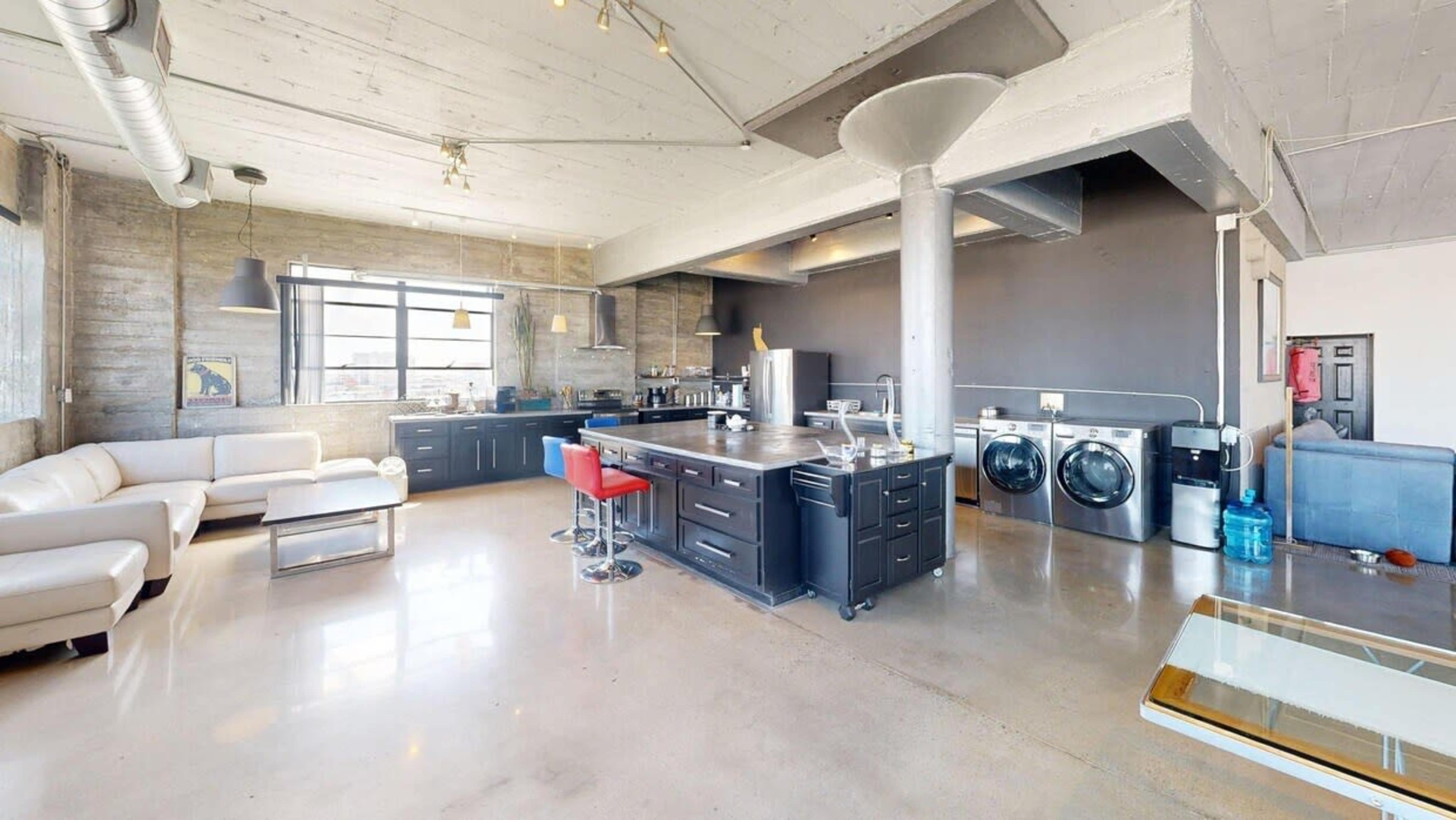 The image features a modern, open-concept kitchen and living area with a concrete floor, stainless steel appliances, and a white leather couch.