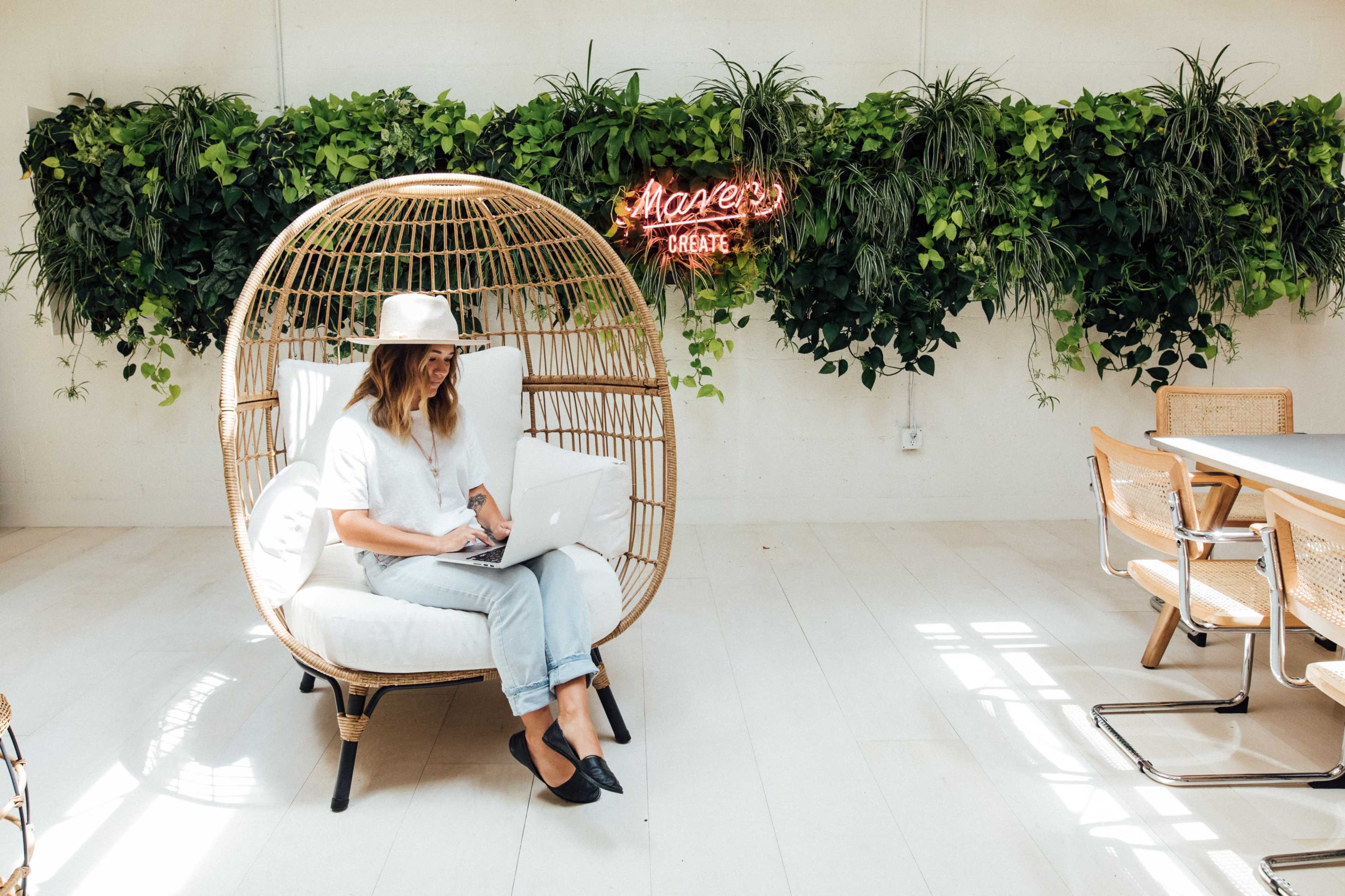 A woman sits in a large wicker chair, working on a laptop in a bright room adorned with hanging plants and a neon sign that says "Create."