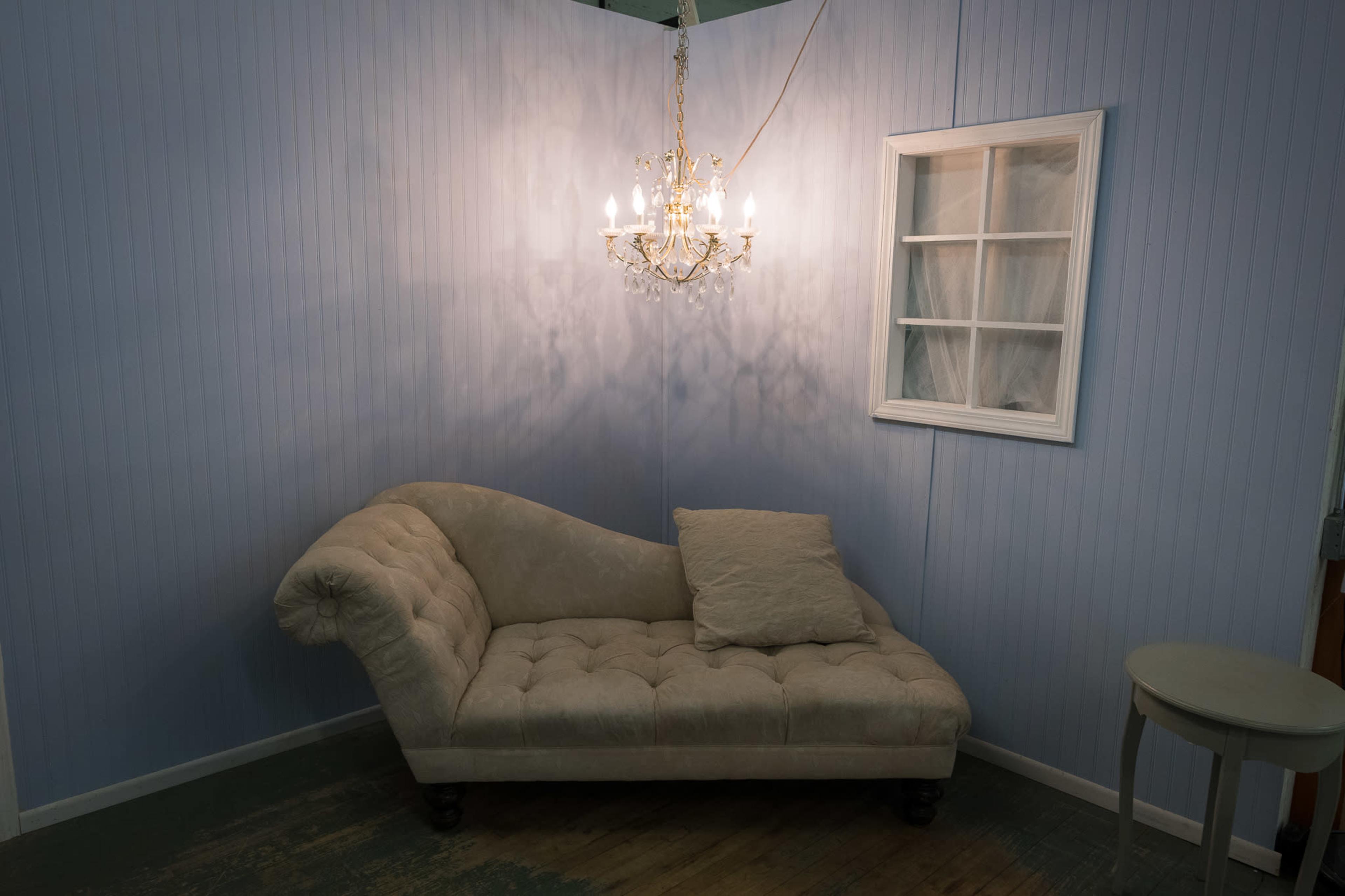The image shows a lightly colored chaise lounge with a throw pillow, positioned near a window and a chandelier on the wall in a softly lit corner of a room.